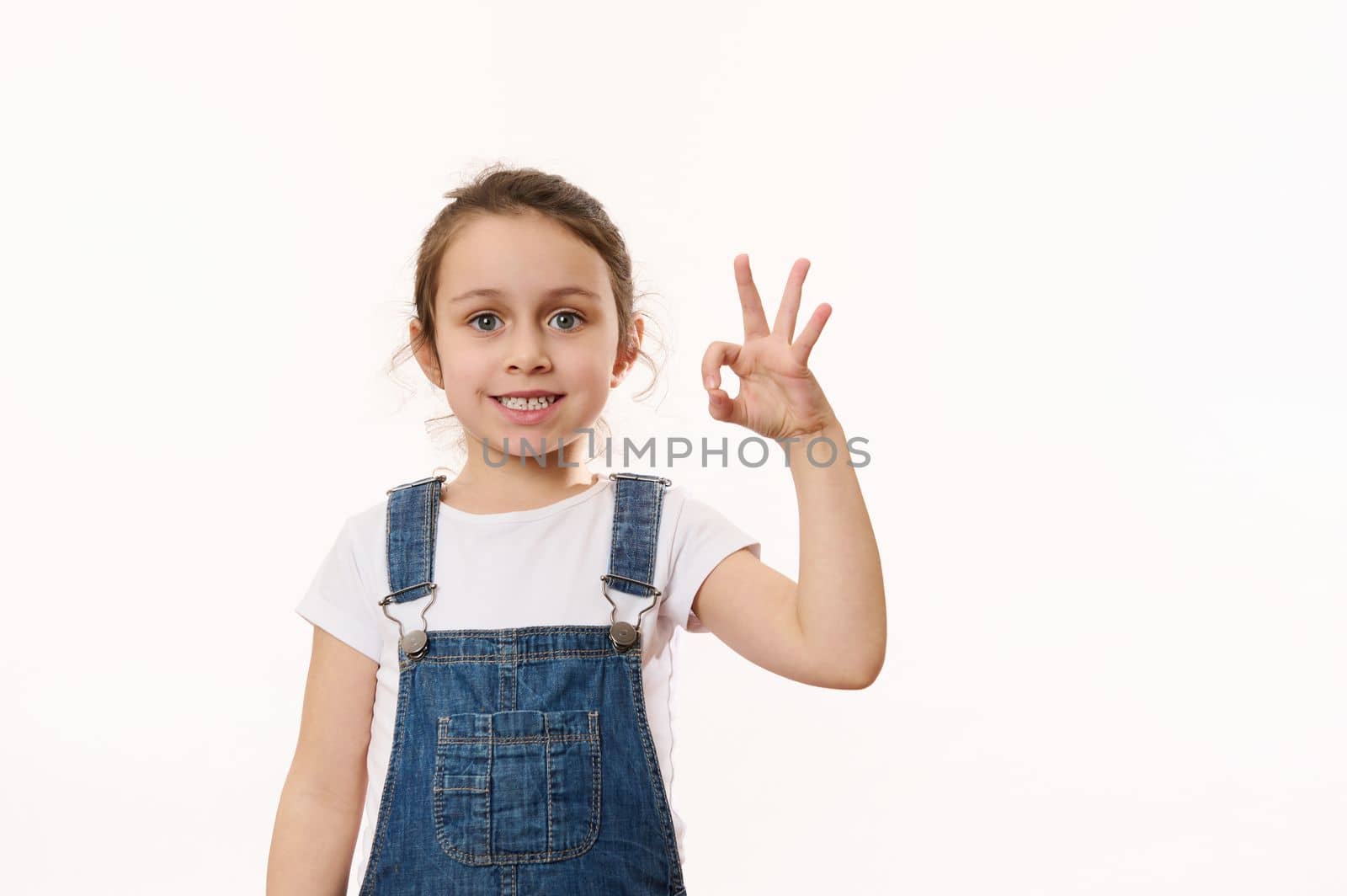 Adorable elementary age, preschool little child girl, in blue denim overalls, gestures, shows peace sign, cutely smiling cheerful toothy smile, looking at camera, isolated white background. Copy space