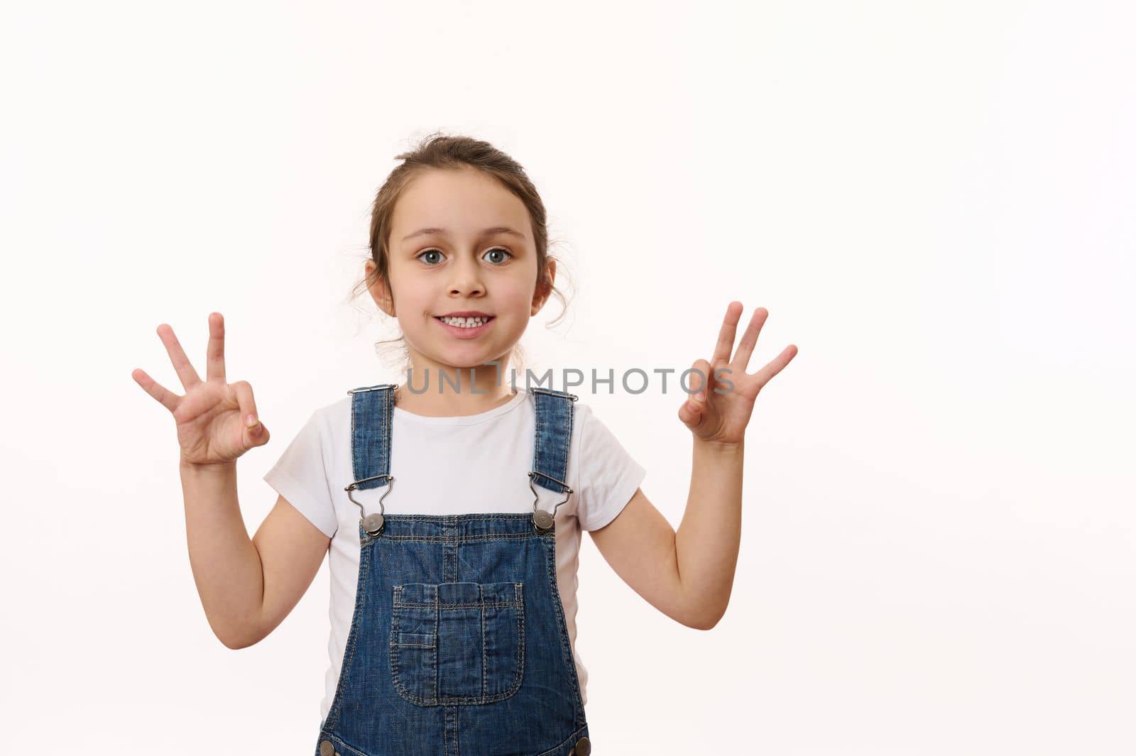 Adorable Caucasian little child, preschool kid, lovely baby girl wearing blue denim overalls, gestures, shows OK signs and happily smiles a toothy smile looking at camera, isolated on white background