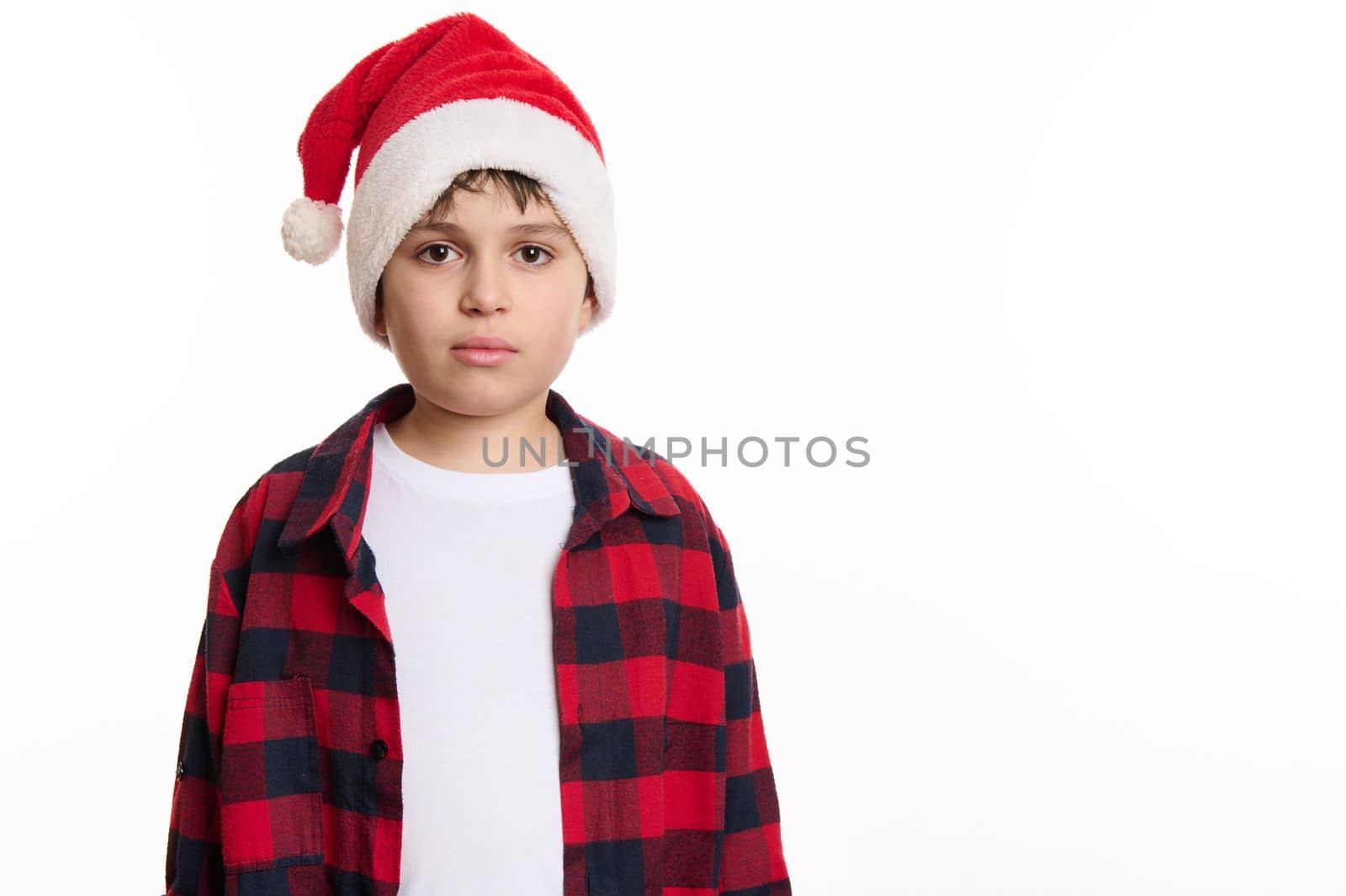 Isolated portrait on white background of a handsome Caucasian preteen boy in Santa hat, in anticipation of Christmas holidays. Pure joy. Winter spirit. New Year party. Copy space for advertising text