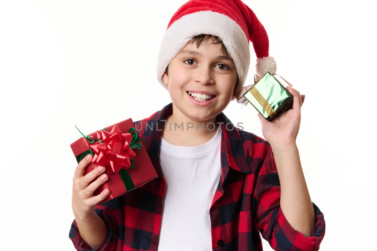 Adorable Hispanic boy wearing Santa hat, smiling a beautiful toothy smile, posing with Christmas gifts, looking at camera. New Years preparations. Winter holidays, magical festive atmospheric mood