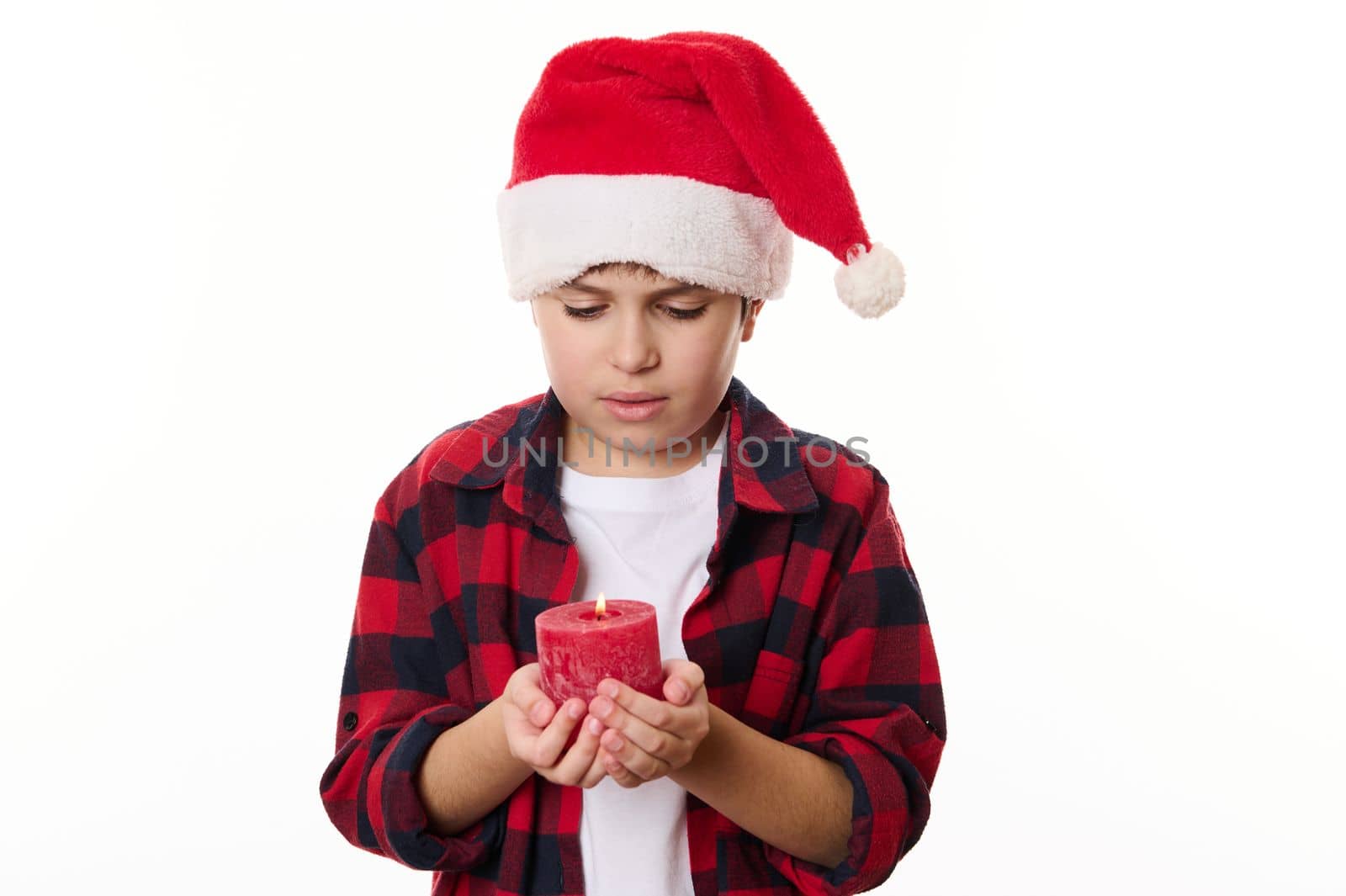 Charming Caucasian child boy in Santa hat, holds a red lit candle, making wish for Christmas, isolated on white background with copy advertising space. Magical atmosphere of winter holidays