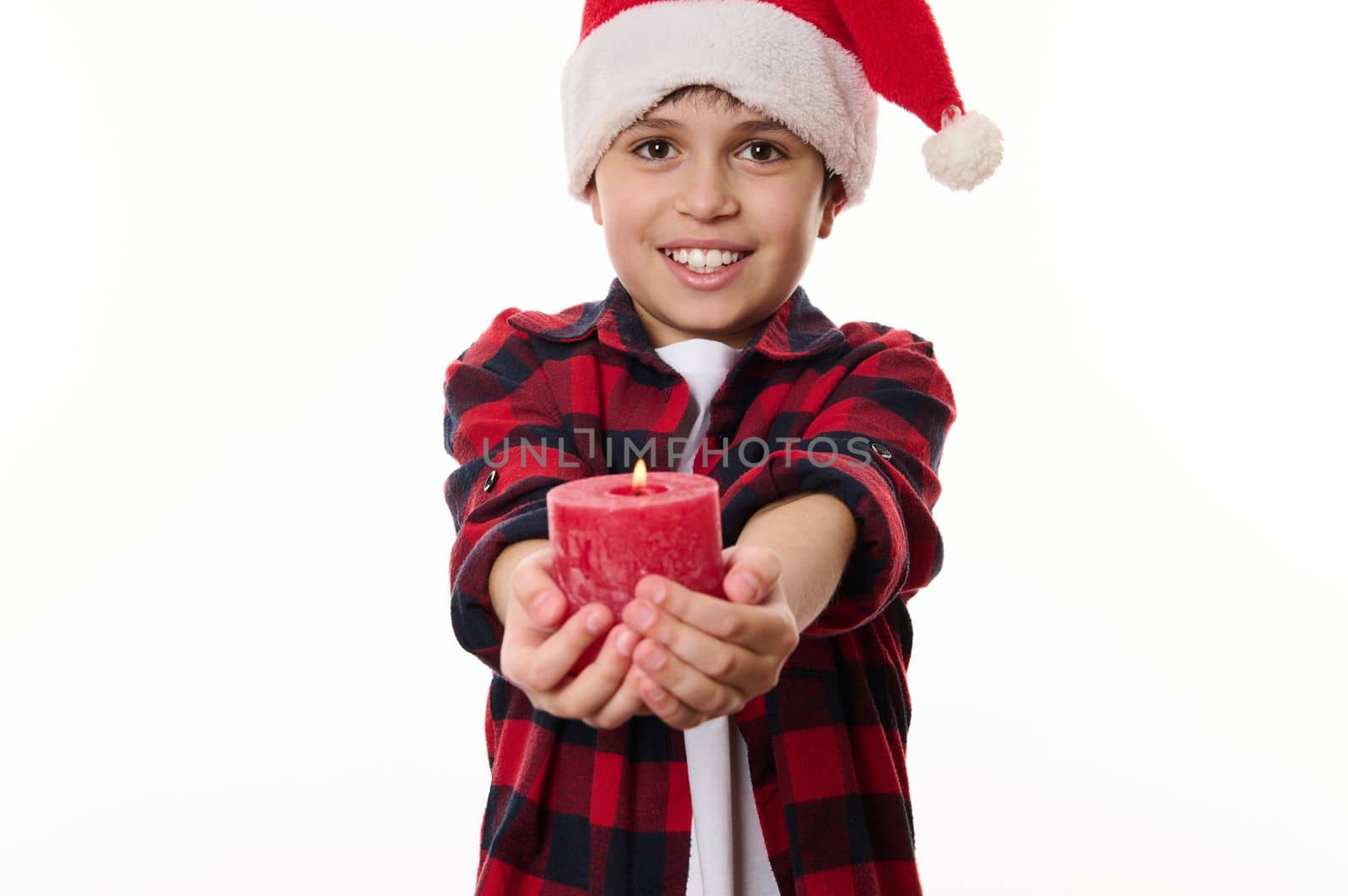 Happy Caucasian teenage boy in Santa hat and red plaid shirt, smiling a beautiful toothy smile , holding a lit Christmas candle, looking at camera. Isolated on white background. Copy advertising space