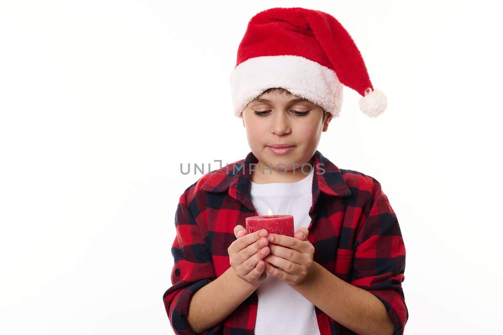 Close-up portrait of handsome Caucasian teenage boy wearing Santa hat and red plaid shirt, making a cherished wish while holding a lit Christmas candle. Isolated on white background. Copy ad space