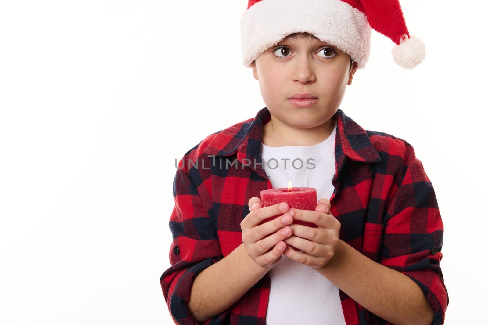 Adorable thoughtful Caucasian preteen boy in Santa hat and plaid shirt, with a red lit Christmas candle, isolated on white background with copy advertising space. Magical atmosphere at winter holidays