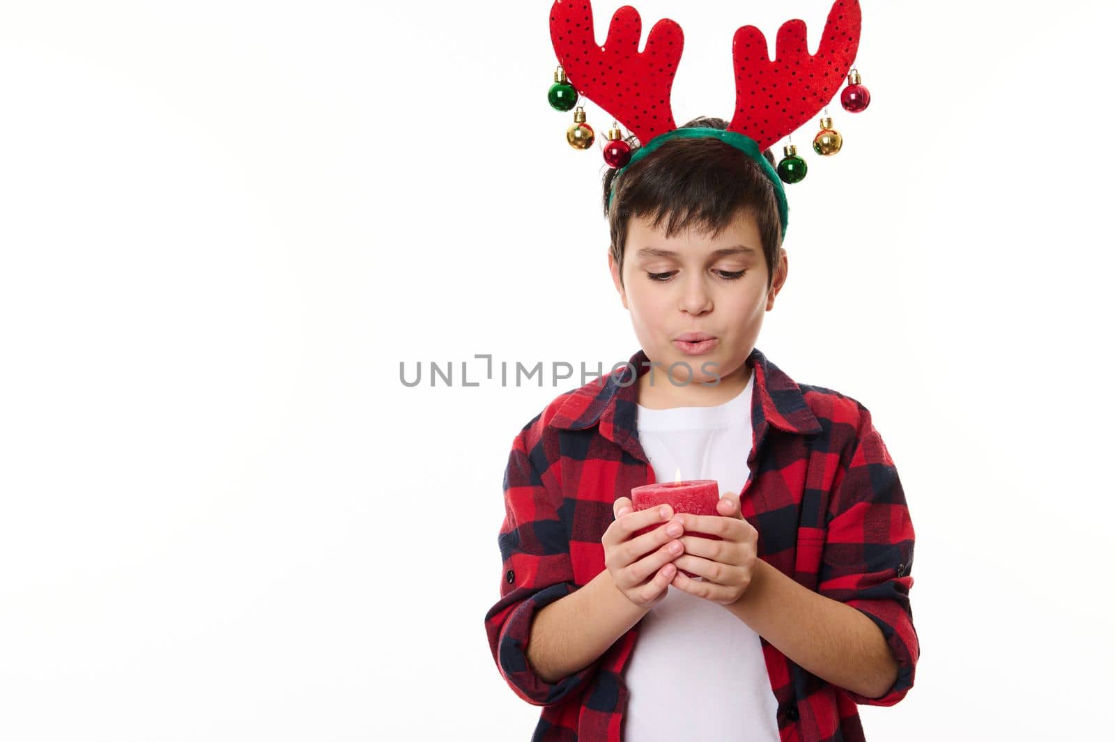 Isolated portrait on white background of an adorable child boy, with deer antler hoop on his head, making wish for Christmas and blowing out a red candle in his hands, Copy advertising space or text