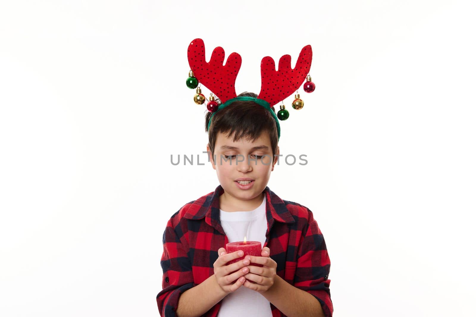 Handsome Caucasian child boy with deer antler hoop on head, holding a decorative lit candle, making cherished wish for Christmas, isolated on white background. Happy childhood. Magical winter holidays