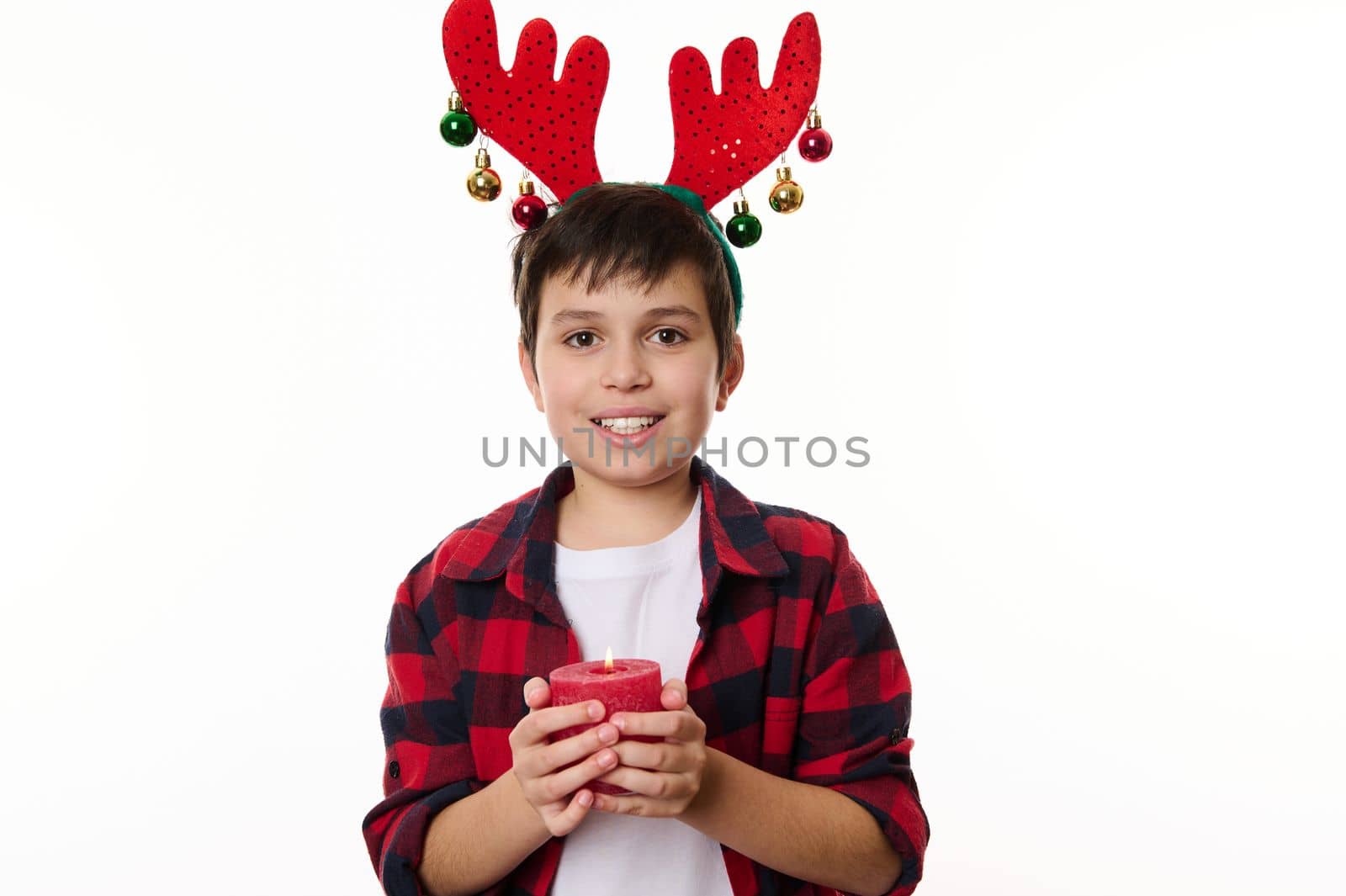 Happy Caucasian teenage boy with deer antler hoop on his head, smiling a beautiful toothy smile, holding a lit Christmas candle, looking at camera. Isolated on white background. Copy advertising space
