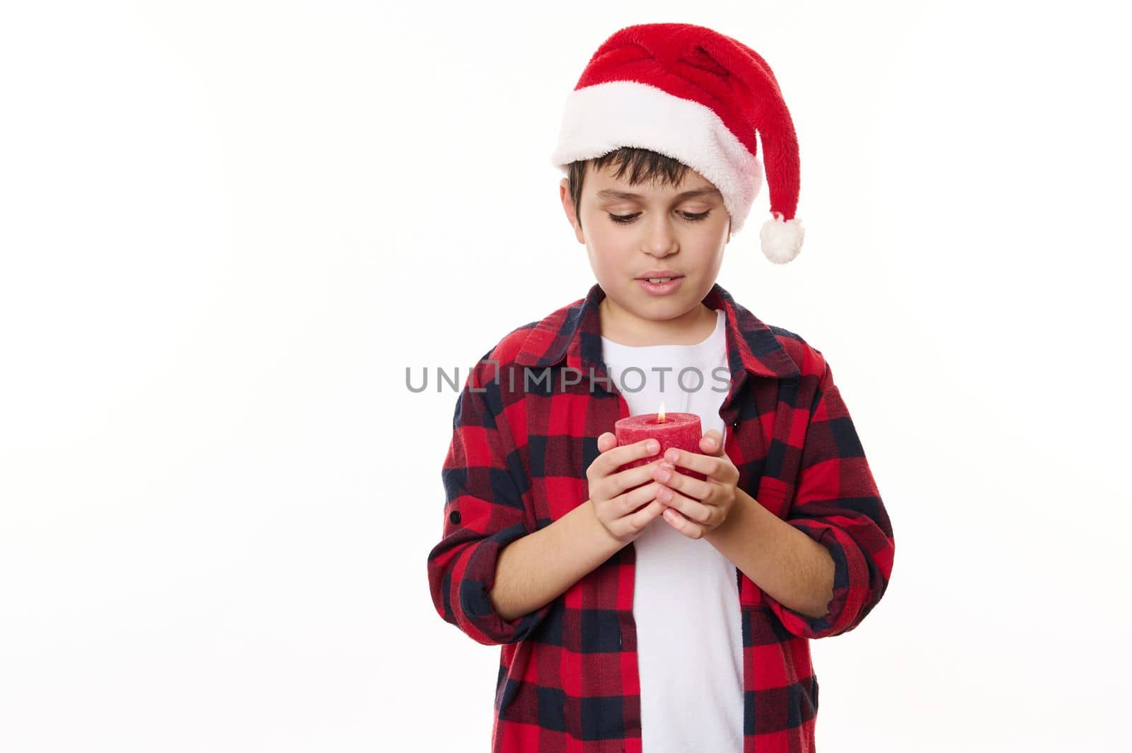 Handsome preteen boy in red checkered shirt and Santa hat, holding a decorative lit candle, making cherished wish for Christmas, isolated on white background. Happy childhood. Magical winter holidays