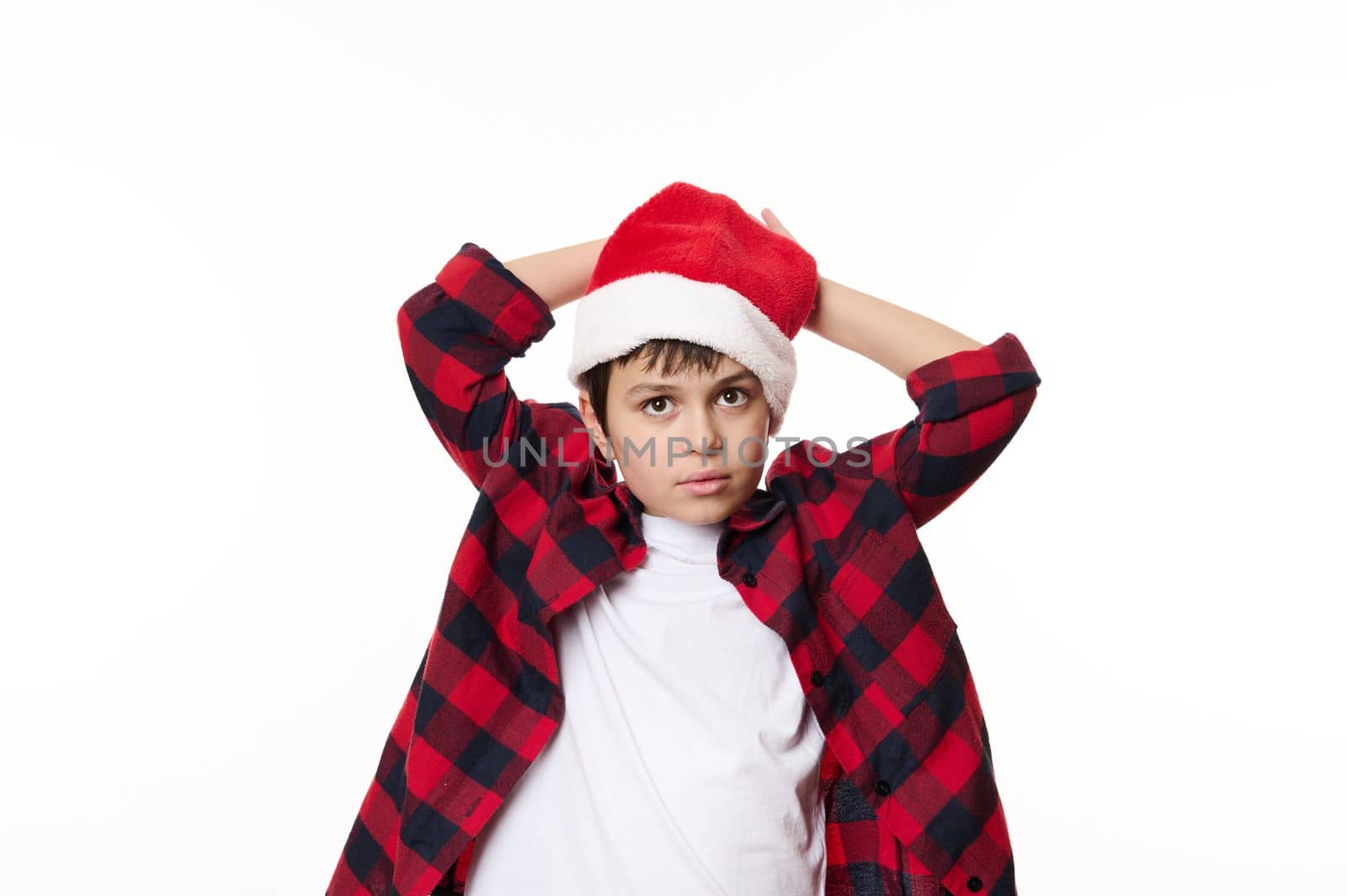 Isolated portrait of a confident handsome pensive preteen boy, wearing Santa hat and red plaid shirt posing over a white background, looking at camera. Ad space. Mmerry Christmas. Xmas. Magic winter