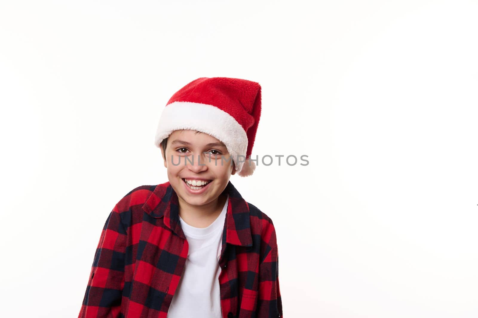Isolated portrait on white background, with copy advertising space, of a charming teenage boy in red Santa hat, laughing and smiling a cheerful toothy smile looking at camera. Facial expressions