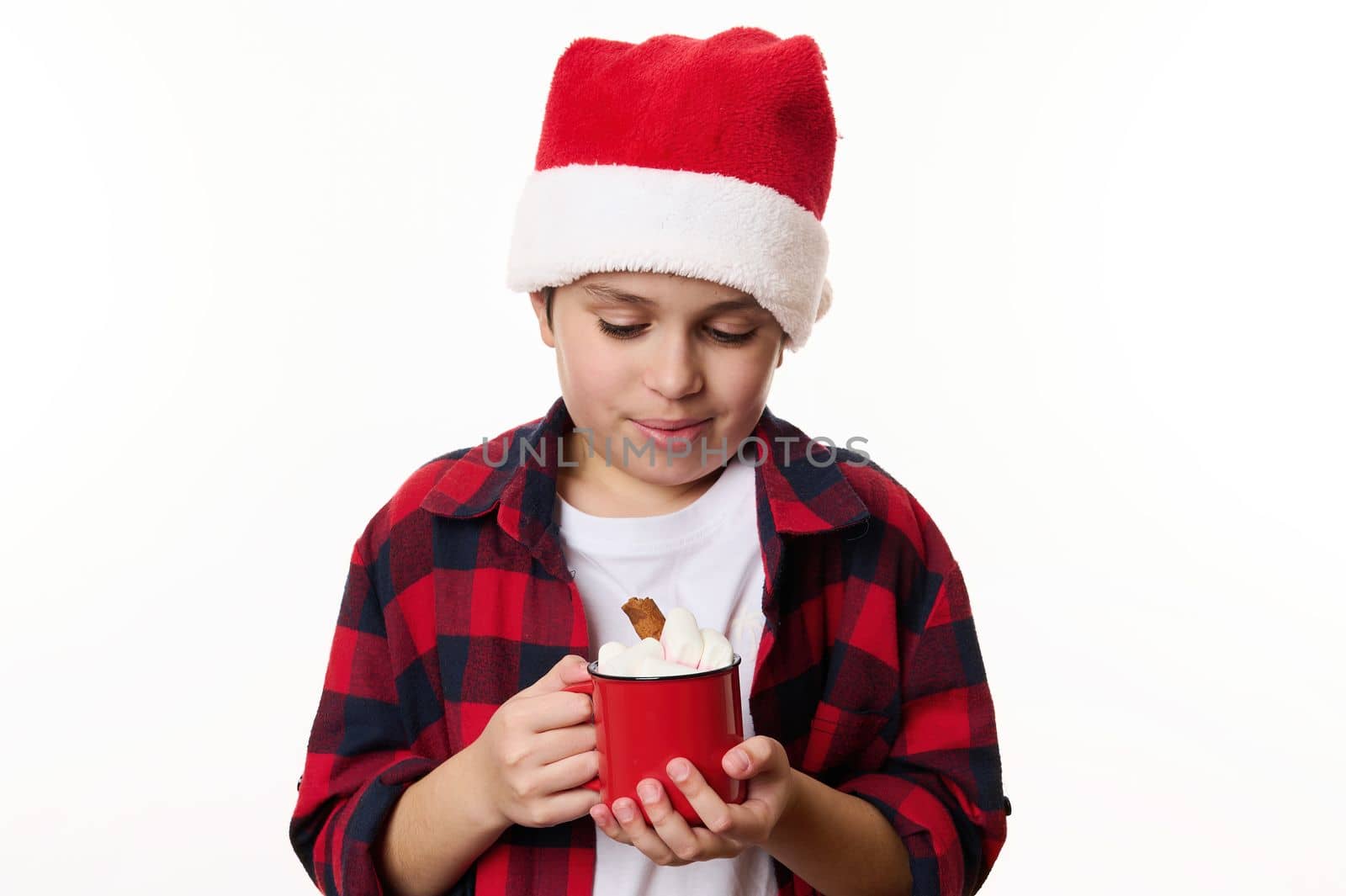 Adorable preteen boy wearing Santa hat, warming his hands with a red cup of hot chocolat and marshmallows, isolated on white background with copy advertising space. Merry Christmas, New Year concept