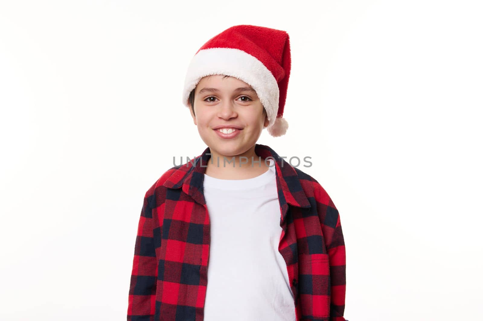 Portrait on white background of adorable teenage boy, wearing Santa hat and red plaid shirt, smiling beautiful toothy smile, looking at camera, feeling happy and anticipating upcoming winter holidays