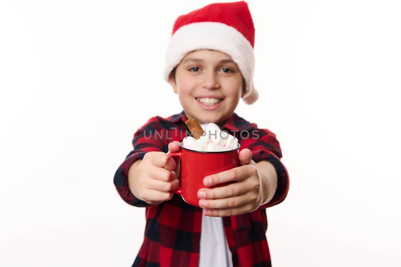 Details: a cup of hot chocolate with marshmallows, in the hands of a blurred cheerful Caucasian school boy in Santa hat, smiling to the camera, isolated on white background with copy advertising space