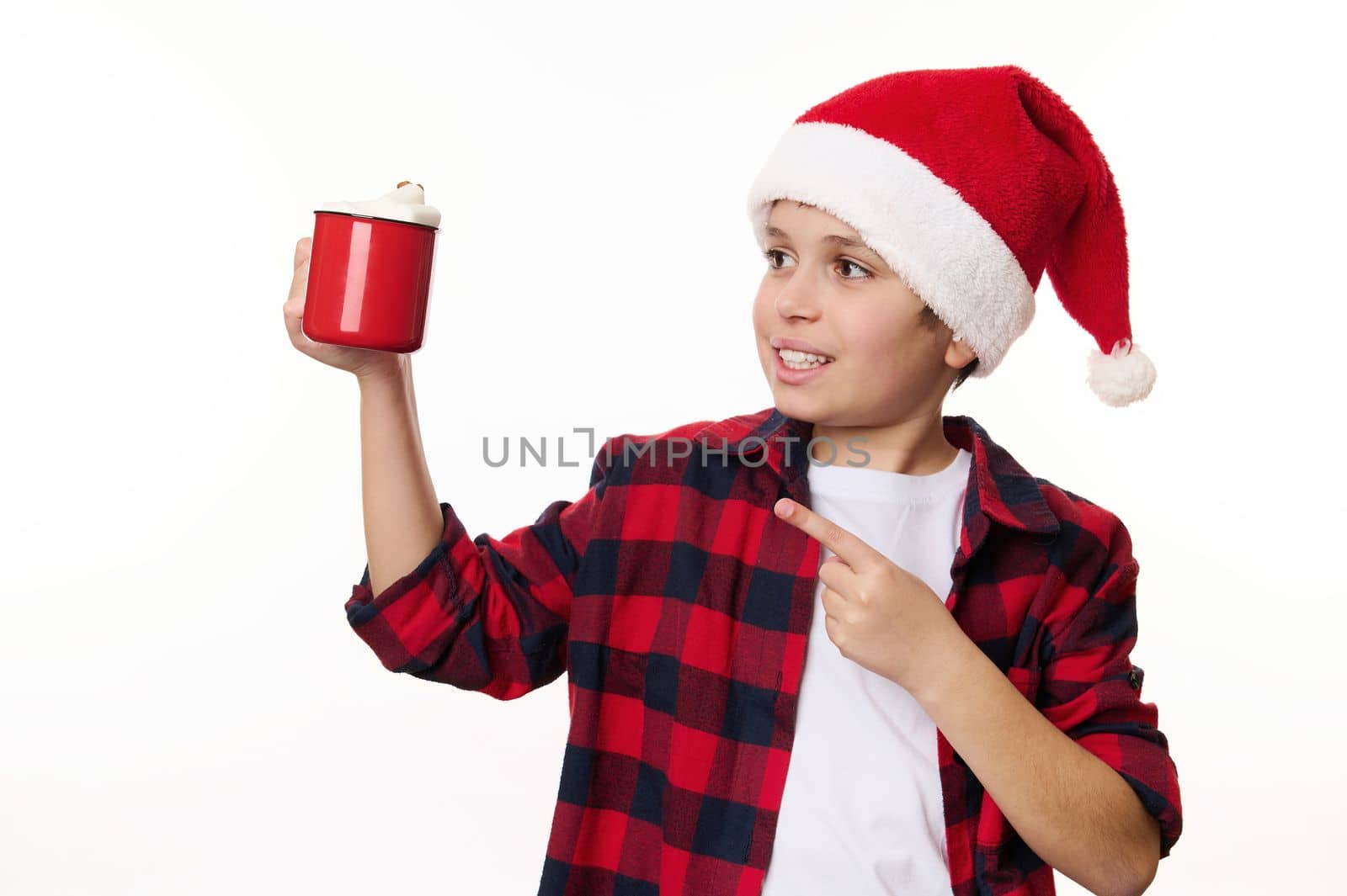 Handsome multi-ethnic preteen boy in Santa hat, pointing on red cup of hot cocoa with marshmallows, isolated on white background. Copy space or promotional text. Christmastime. Happy winter holidays