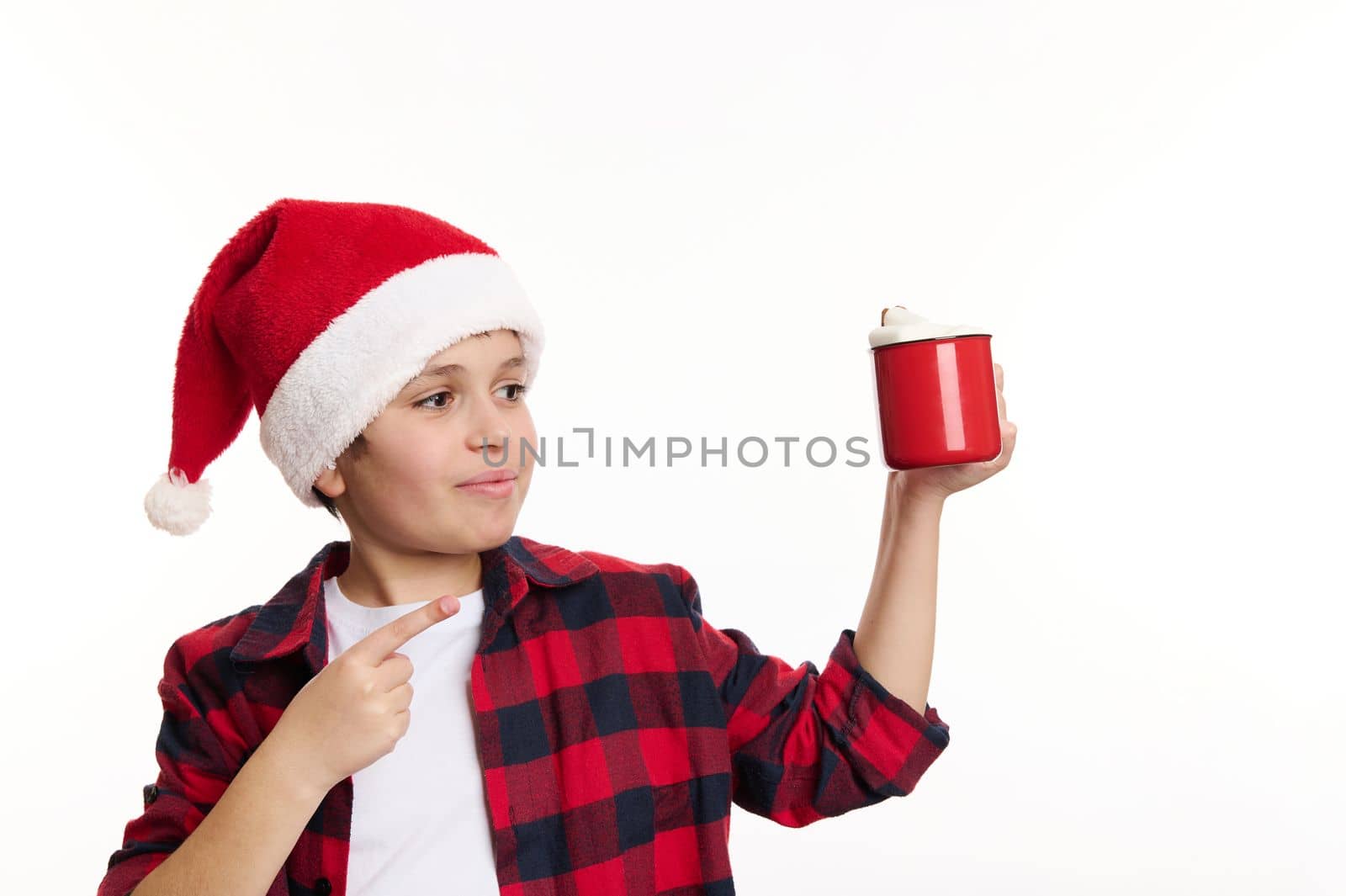 Adorable Caucasian preteen boy in Santa hat, pointing on red mug of hot chocolate with marshmallows, isolated on white background. Copy space or promotional text. Christmastime. Happy winter holidays
