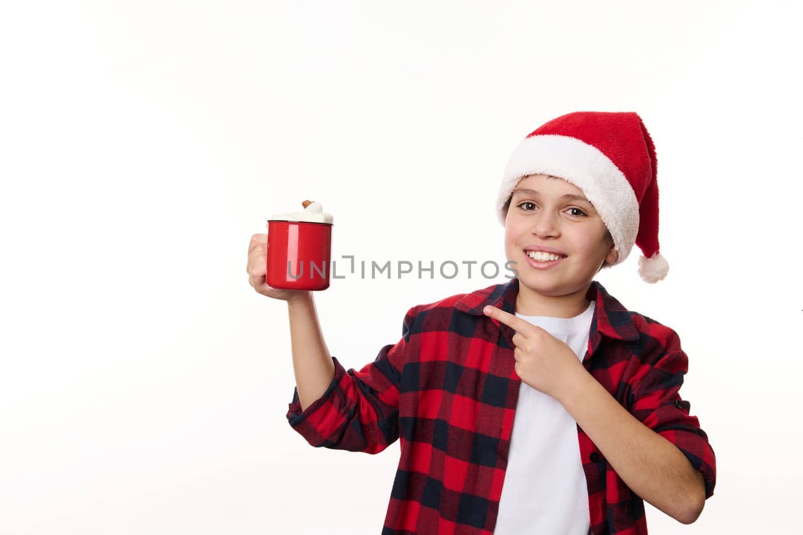 Cheerful child, delightful boy wearing red plaid shirt and Santa hat, with a cup of hot cocoa and marshmallows, smiling a toothy smile looking at camera, isolated on white. Christmas atmospheric mood
