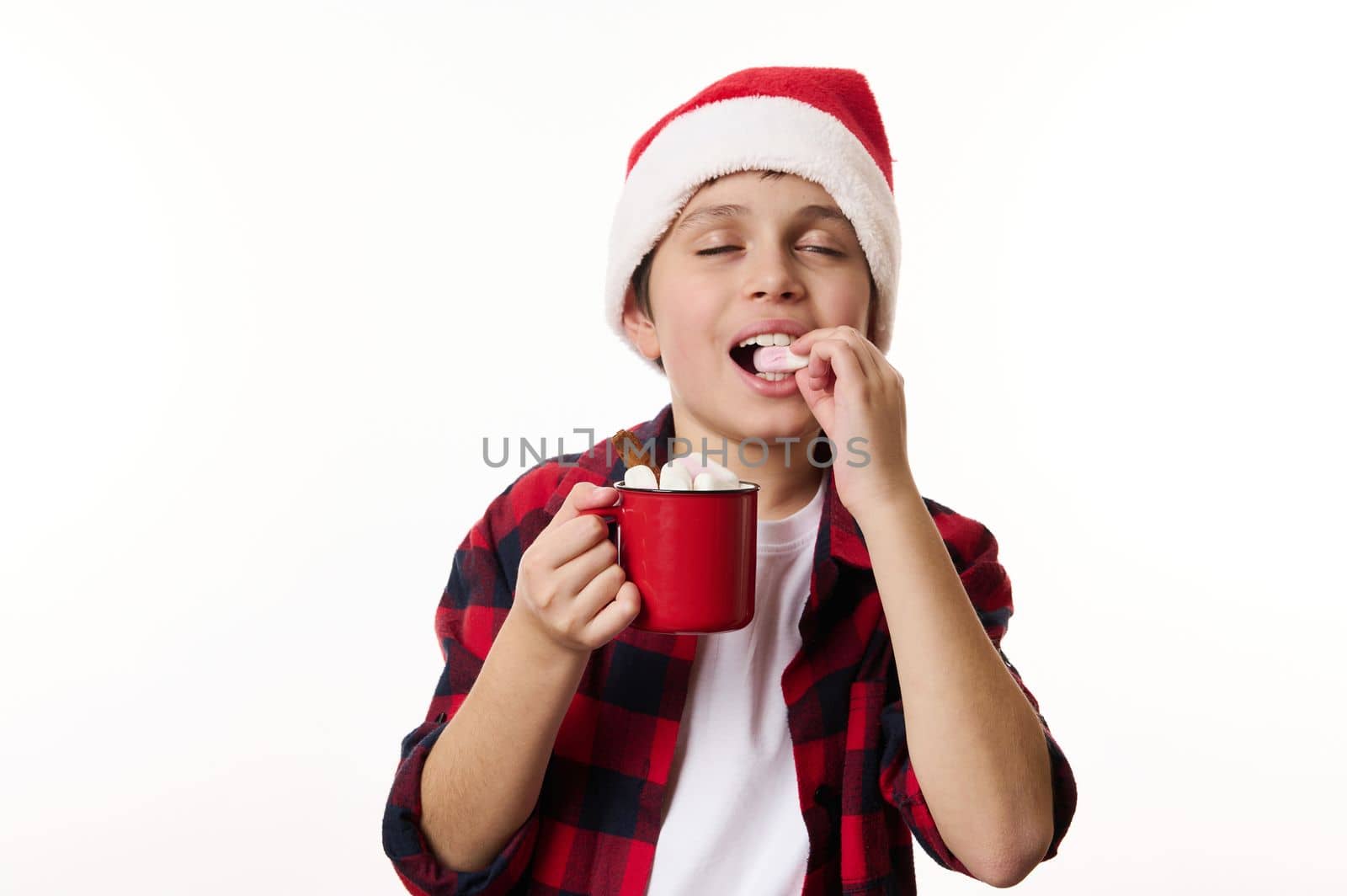 Adorable, delightful child, cheerful boy, teenager in Santa hat, enjoys a hot chocolate drink, eats delicious sweet marshmallows on white background. Copy space. Christmas and winter holidays concept