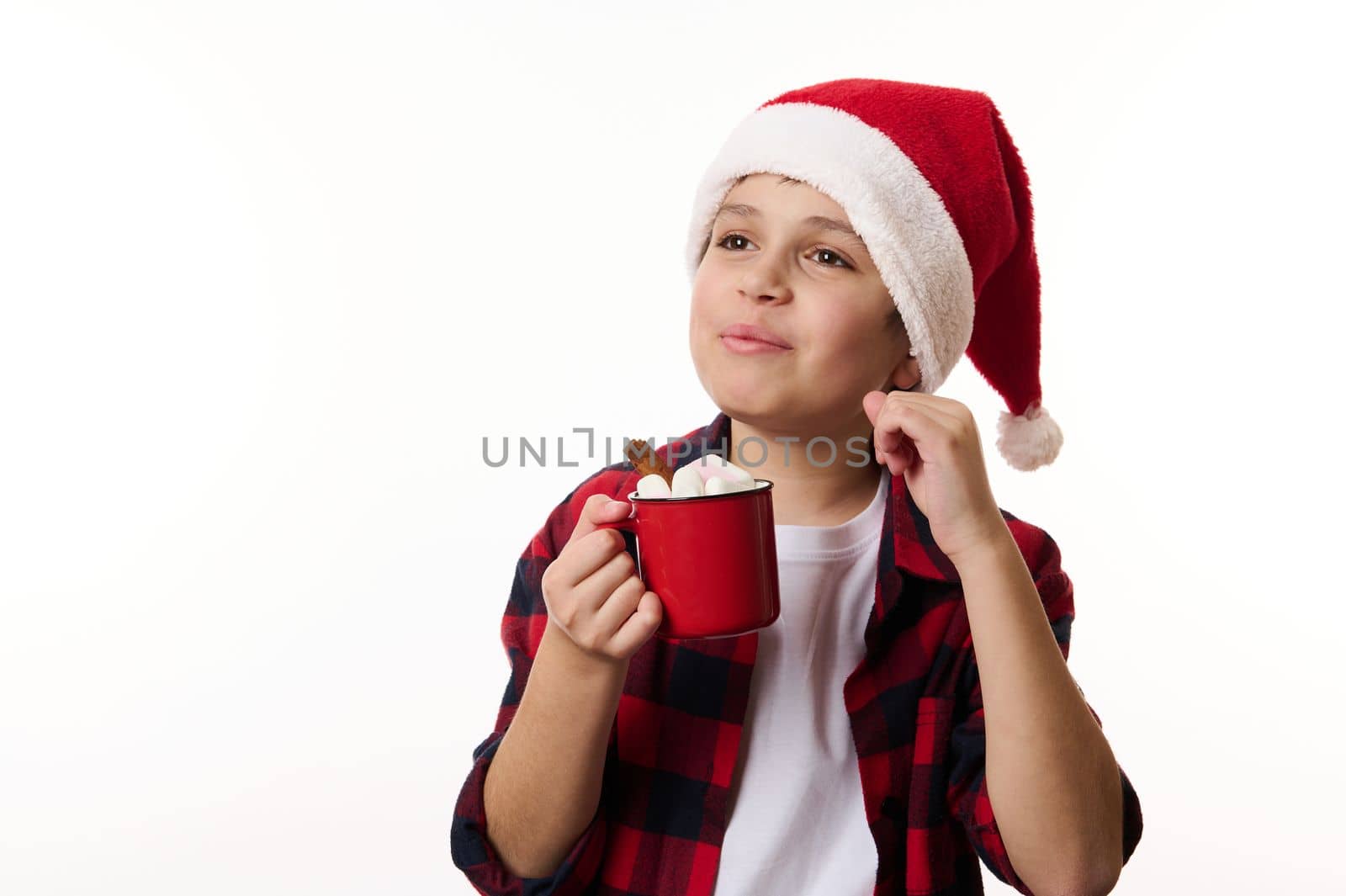 Isolated portrait on white background of a happy boy in Santa hat, enjoys hot chocolate drink with sweet marshmallows. Concept of expecting the upcoming winter holidays. Christmas and New Year's event