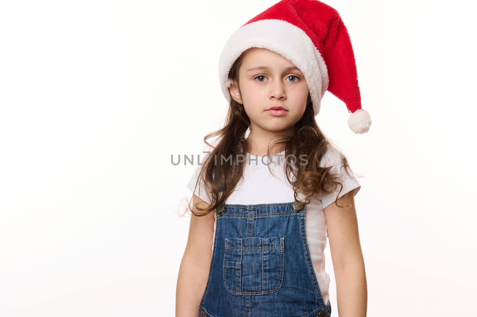 Isolated portrait on white background of a fancy cute Caucasian little girl in santa hat, in anticipation of Christmas holidays. Pure joy. Winter spirit. New Year party. Copy spacefor advertising text