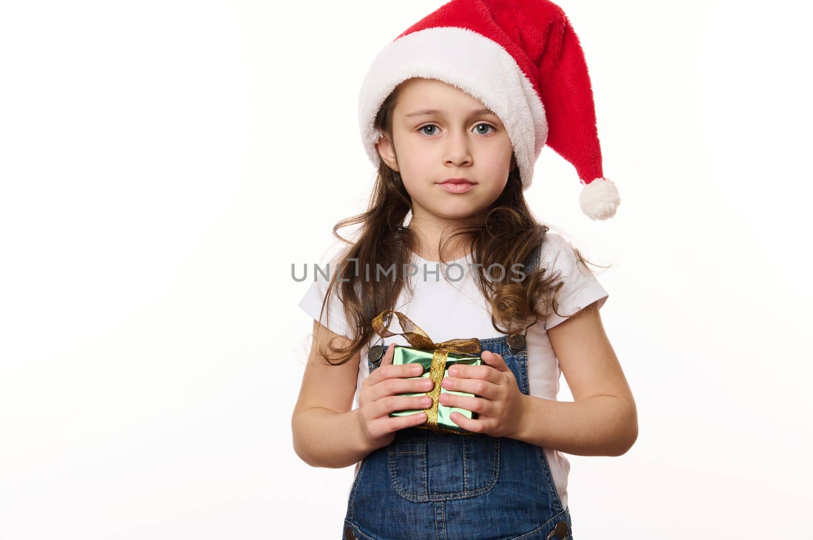 Lovely baby girl, adorable little child wearing Santa hat and blue denim overalls, holds a happy Christmas present wrapped in green shiny gift paper, isolated on white. Christmas time. Winter holidays