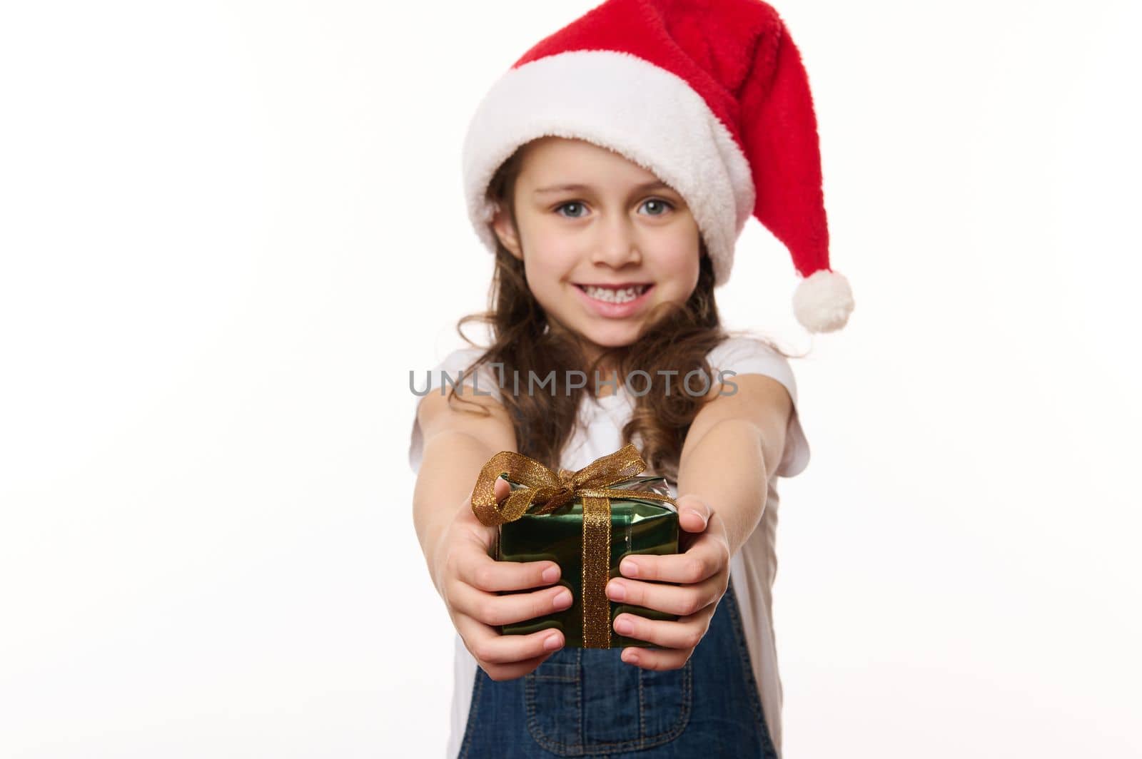 Selective focus on hands of a blurred lovely little girl in Santa hat, holding a cute Christmas present, isolated on white background. Copy space. Christmas Eve. New Year and winter holidays concept