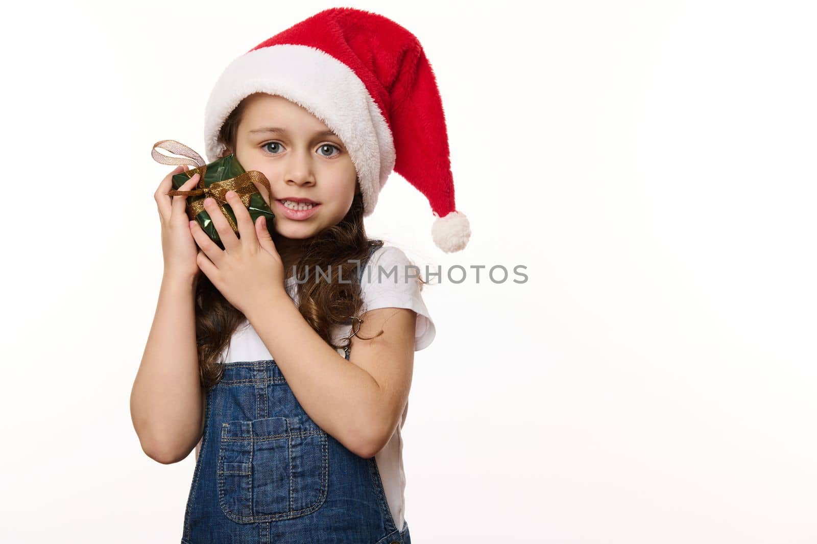 Isolated portrait with copy ad space on white background, of an adorable European little girl wearing santa hat, tenderly hugging her Christmas present. Looking forward to the upcoming winter holidays