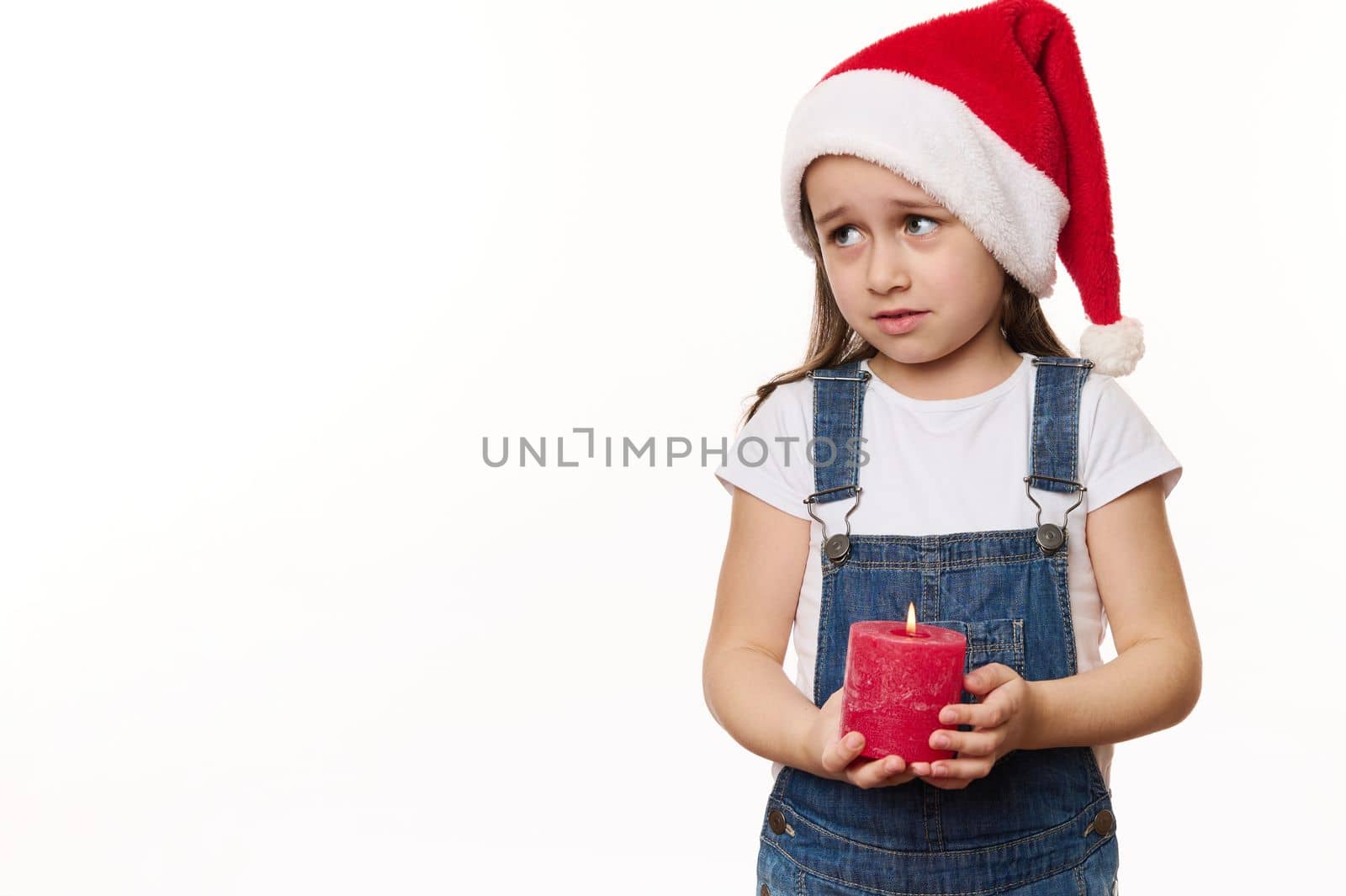 Mischievous little girl in Santa hat, holds a lit candle, makes wish, looks at copy ad space for text on white background. Looking forward to upcoming winter holidays. Merry Christmas. Happy New Year.