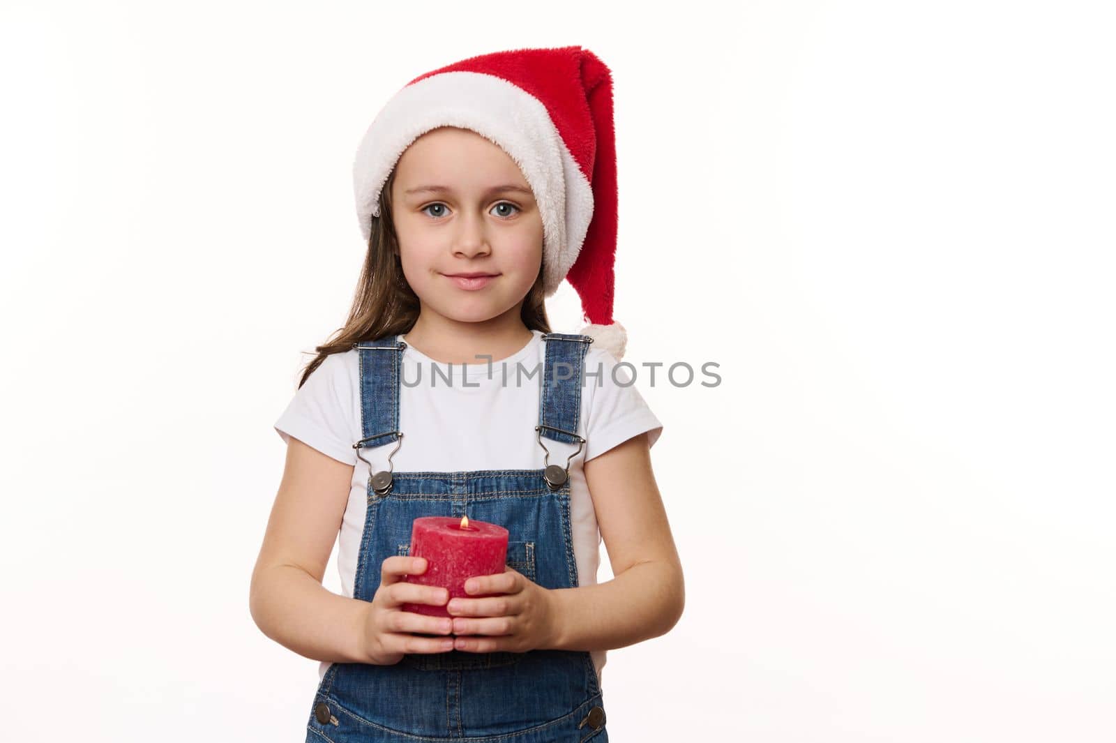 Delightful preschooler girl, lovely little child in Santa hat and blue denim overalls, holds a lit red candle in her hands and cutely smiles looking at camera, isolated on white background. Christmas
