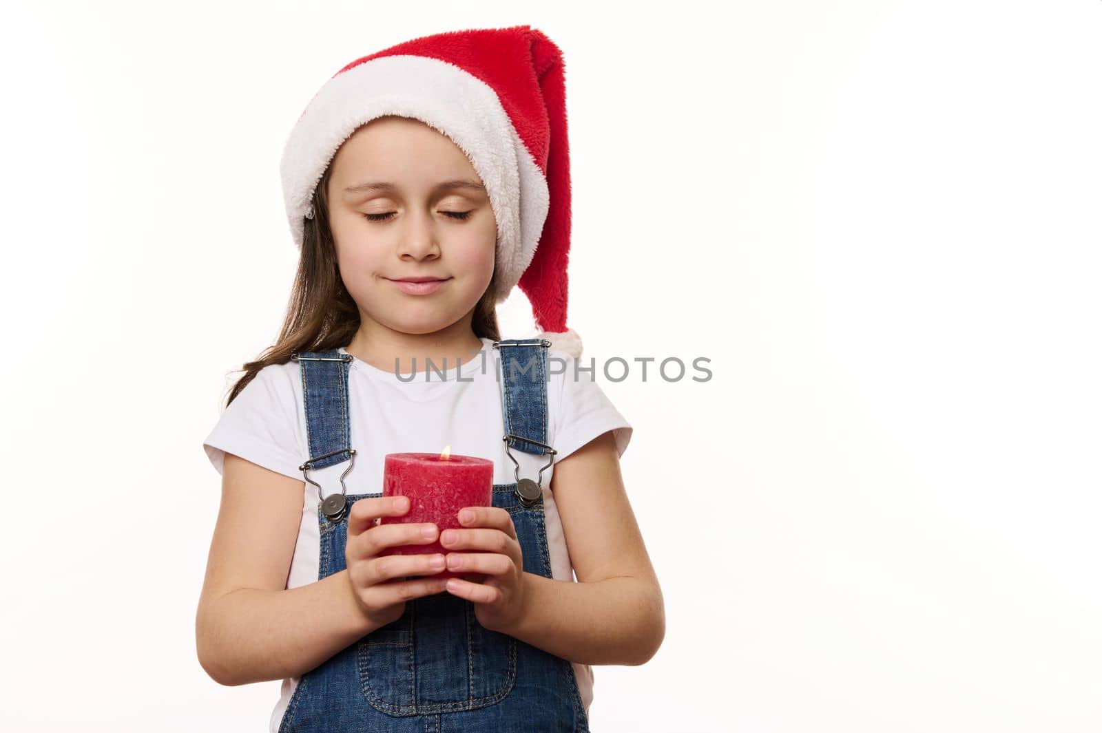 Adorable peaceful little girl in Santa hat, holds a lit candle, makes cherished wish while posing with her eyes closed, against a white background. Copy space. Christmas Eve. New Year. Winter holidays