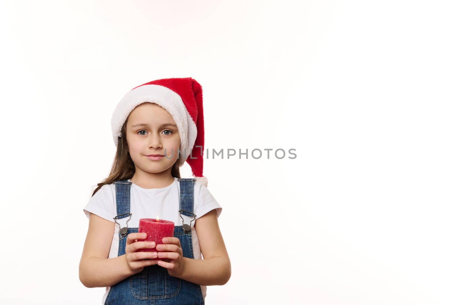 Adorable European baby girl in Santa hat, blue denim overalls, with a lit red candle in hands, isolated over white background. Cute kid makes cherished wish for Christmas. Winter holidays. Copy space