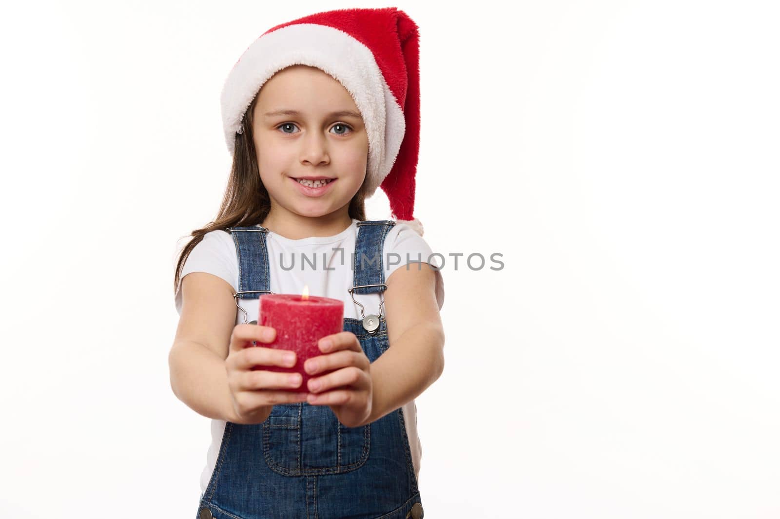 Adorable European baby girl in Santa hat, blue denim overalls, with a lit red candle in hands, isolated over white background. Cute kid makes cherished wish for Christmas. Winter holidays. Copy space
