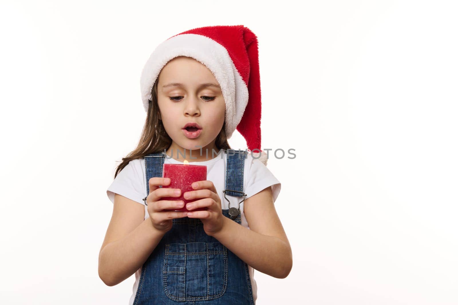 Adorable Caucasian little child, cute baby girl in Santa hat, makes cherished wish for Christmas, blows out a candle, isolated on white background. Happy childhood. Magic atmosphere at winter holidays