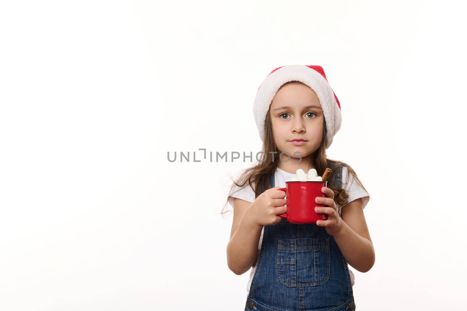 Isolated portrait on white background of a happy little girl in Santa hat, enjoying a delicious sweet hot chocolate drink with marshmallows. Christmas. Winter holidays concept. Copy ad space for text