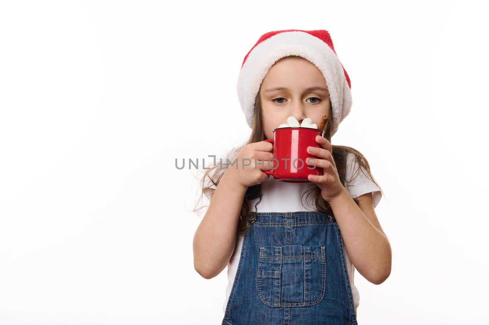 Isolated portrait on white background of a cute baby girl, wearing Santa hat, drinking a delicious hot chocolate with marshmallows. Magical atmosphere of the upcoming Christmas holidays. Celebration