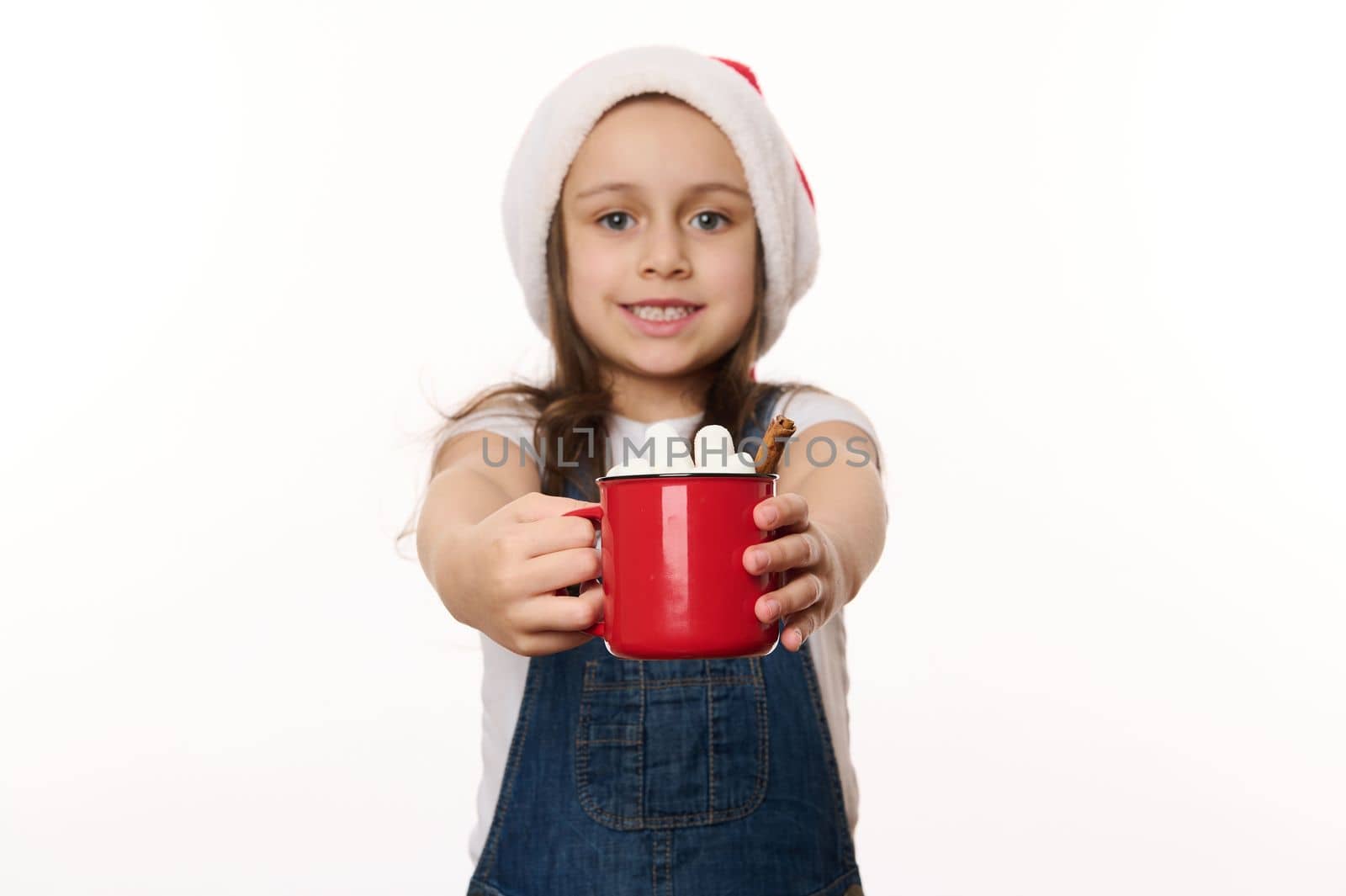Selective focus on a red cup of hot cocoa drink with marshmallows, in the hands of a blurred lovely baby girl wearing Santa hat. Traditional food and beverage for Christmas. Winter festive atmosphere