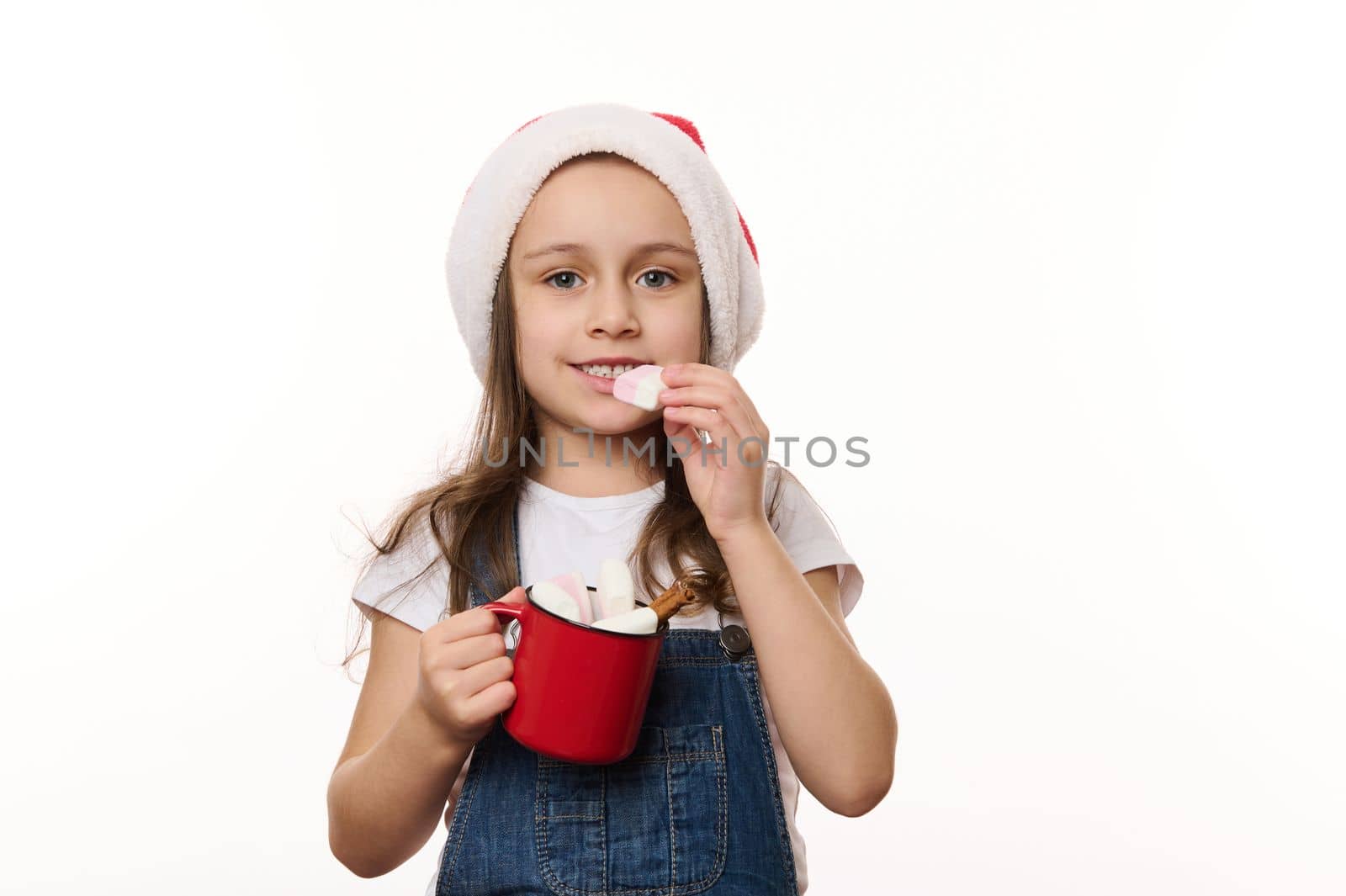 Isolated portrait on white background of lovely little child girl, in Santa hat, holding a red cup of hot chocolate and eating marshmallows. Magic atmosphere of upcoming Christmas, winter holidays.