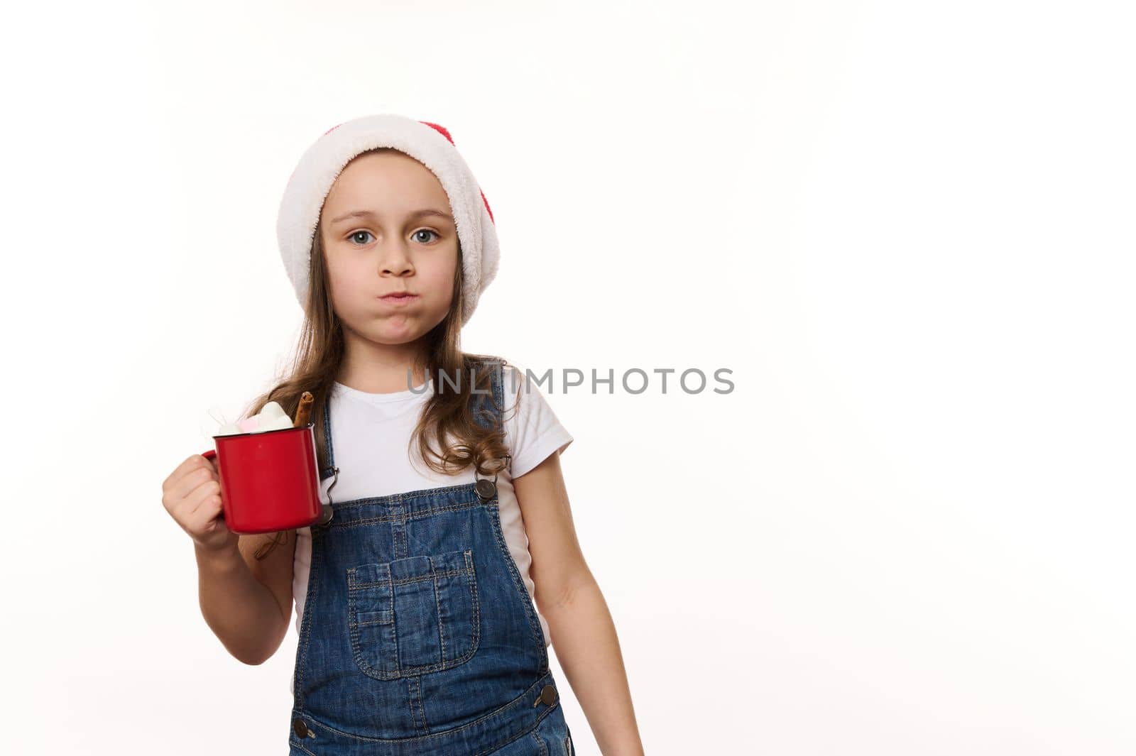 Mischievous 5 years old little girl wearing Santa hat and denim overalls, drinking hot chocolate drink and eating marshmallows, isolated on white background with copy ad space. Christmas atmosphere