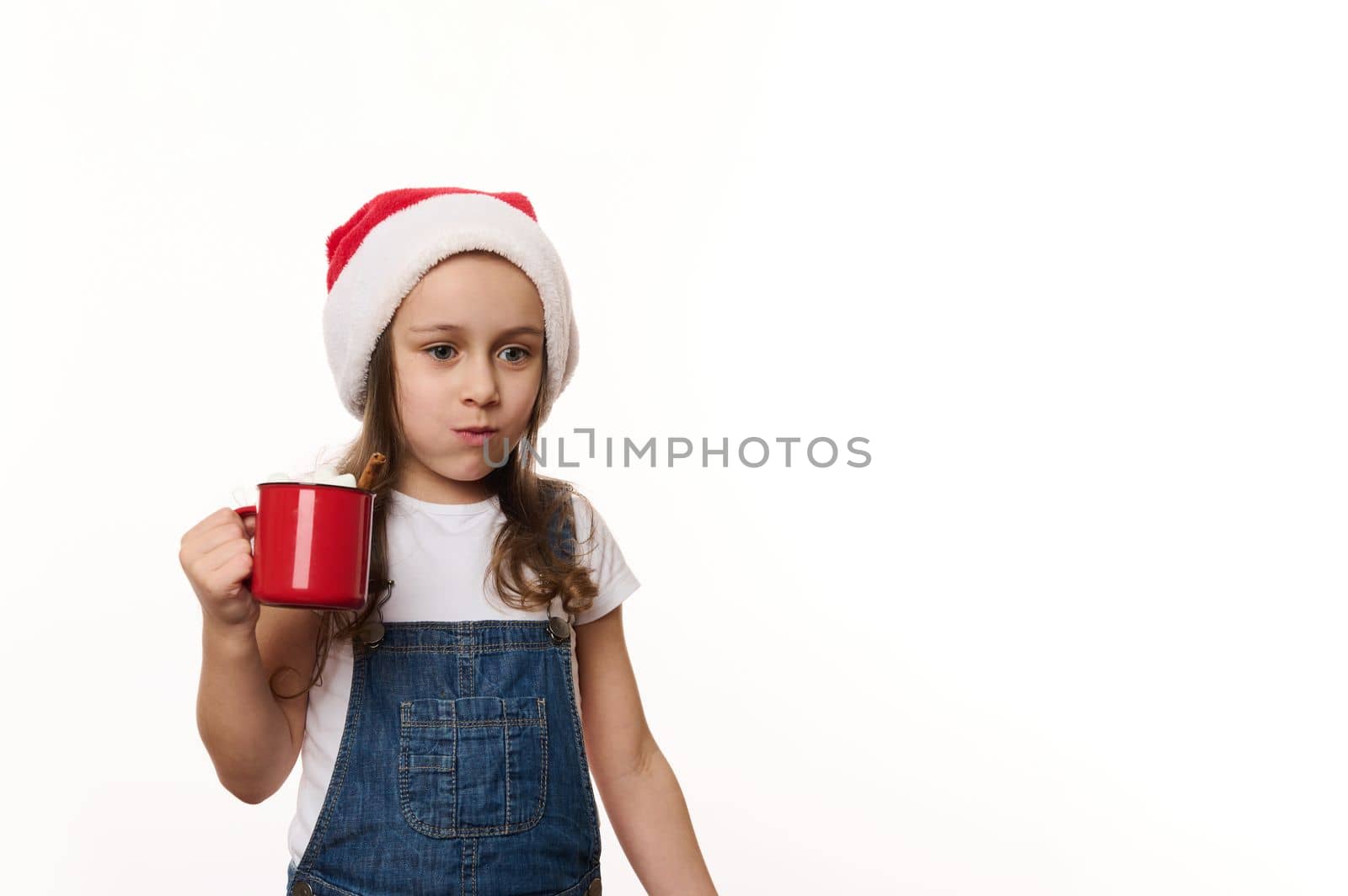 Mischievous lovely baby girl wearing Santa hat and denim overalls, drinking hot cocoa drink and eating sweet marshmallows, isolated on white background with copy ad space. Magic Christmas atmosphere