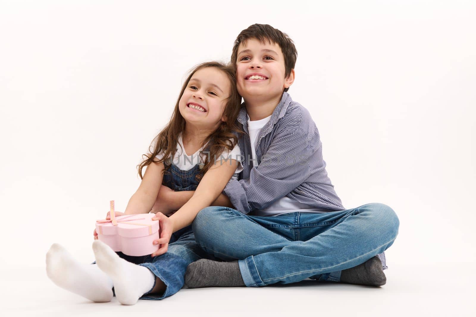 Mischievous adorable kids, a handsome boy and cute girl - a caring brother hugging his younger sister holding a beautiful pink heart-shaped gift box, isolated over white background with copy space