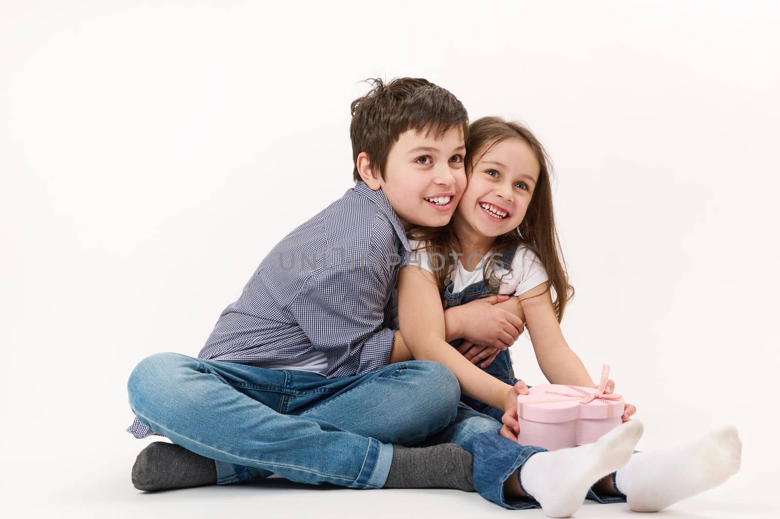 Beautiful kids wearing blue denim jeans and white t-shirt holding a cute heart-shaped gift box, hugging each other, cutely smiling at camera, isolated over white background with copy advertising space