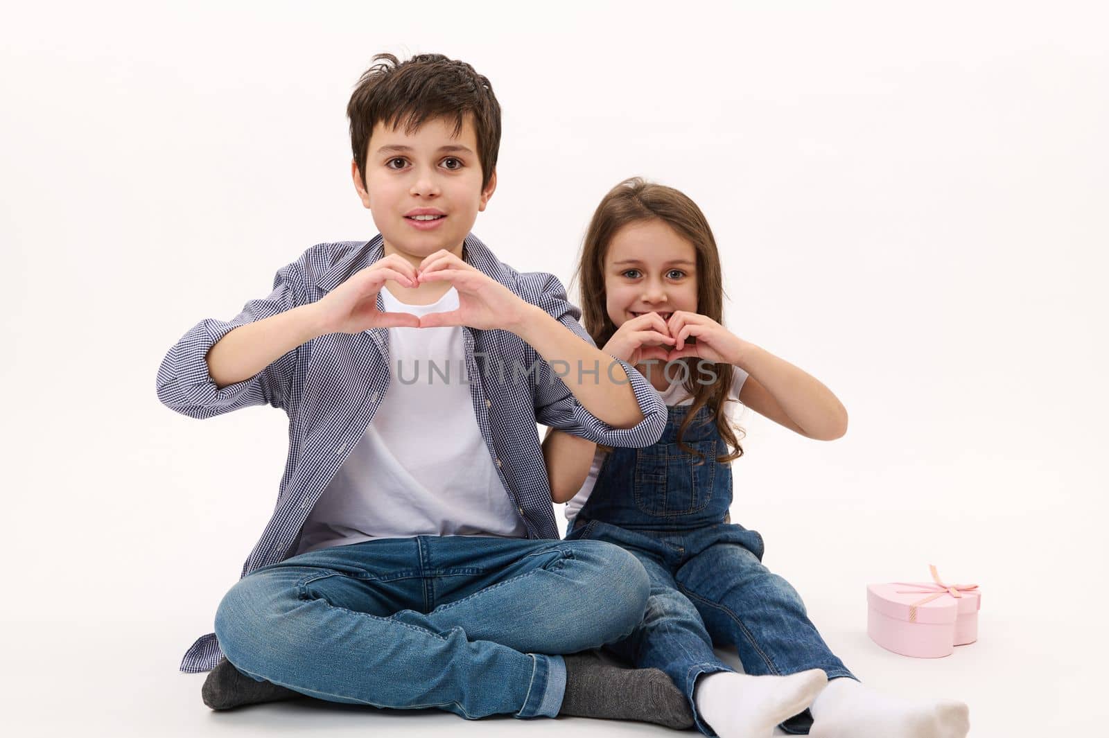 Adorable Caucasian children, boy and girl wearing casual shirt and denim jeans, gesturing showing a heart from fingers, smiling looking at camera, isolated over white background. Saint Valentines Day