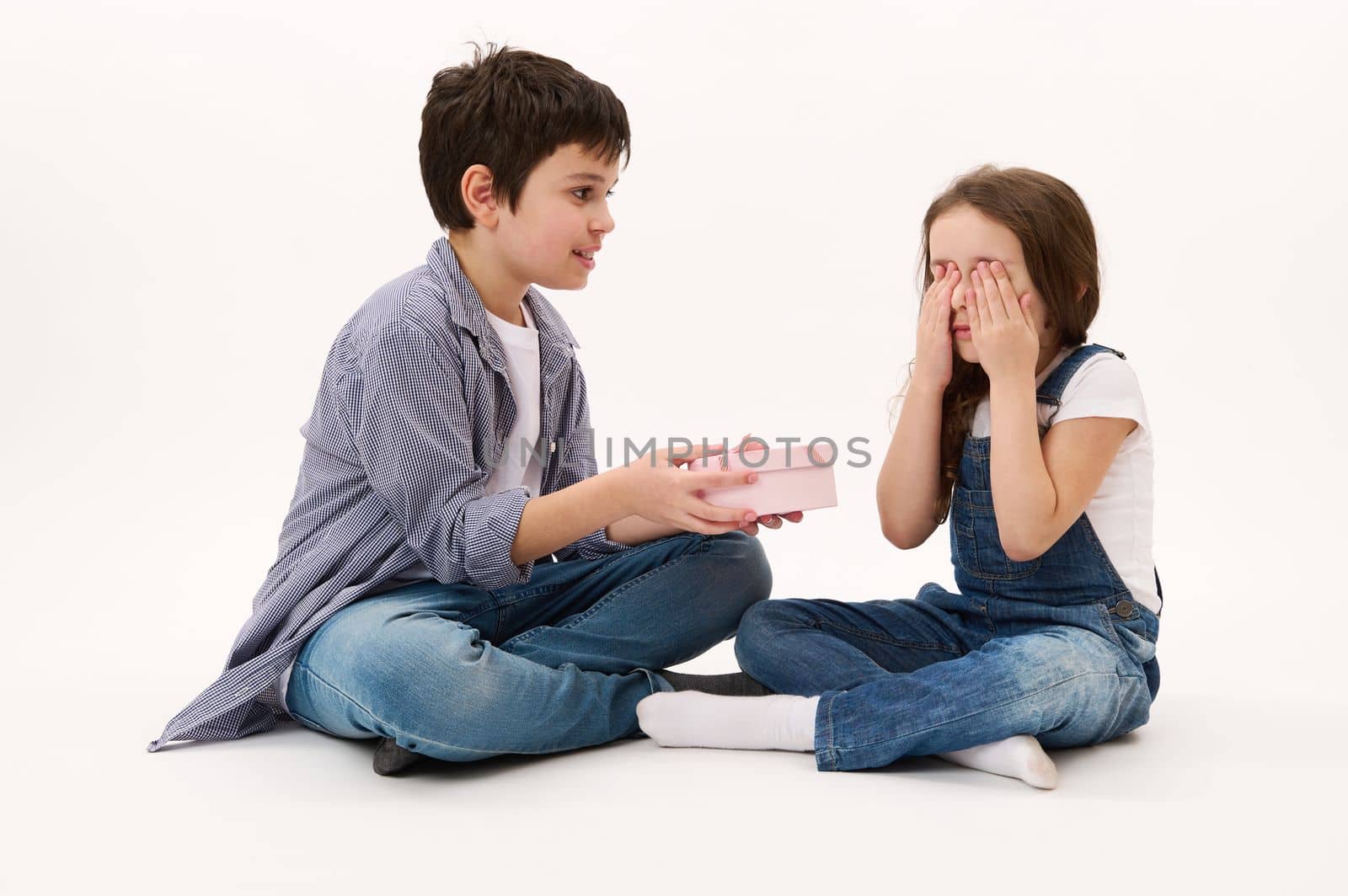 Portrait on white background of adorable teenage boy, loving brother giving a cute present to his younger sister, a little girl covering her eyes. He's greeting her with St Valentine's or women's Day