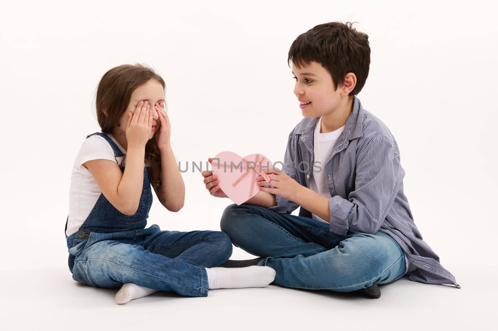 Adorable Hispanic teenage boy, brother gives beautiful heart-shaped pink gift box to his lovely sister, greeting her with St Valentine's Women's Day, isolated on white background. Copy space for text