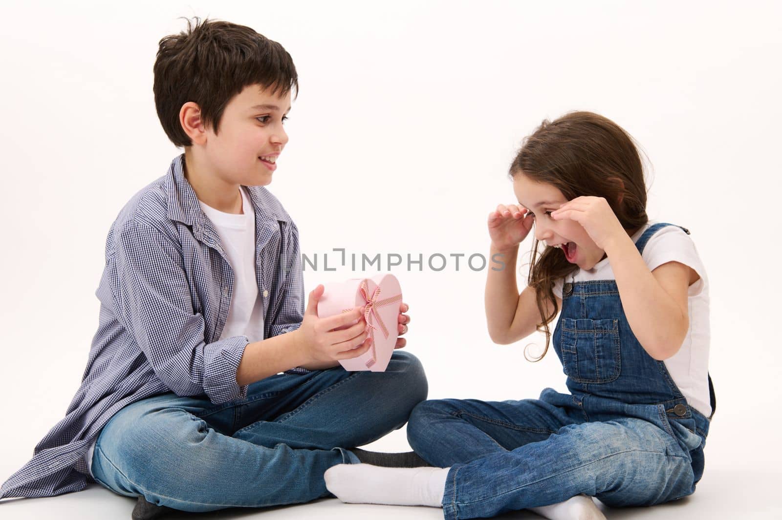 Happy Caucasian little girl in blue denim overalls, smiles cheerfully, rejoices in receiving a cute heart shaped pink gift box from her brother, isolated on white background. Women's Day. March 8th