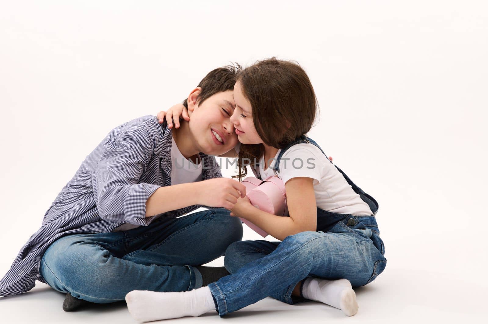 Happy Caucasian little girl in blue denim overalls, rejoices in receiving a cute heart shaped pink gift box from her brother, gently hugging him. Saint Valentines Day. February 14. Women's Day. March