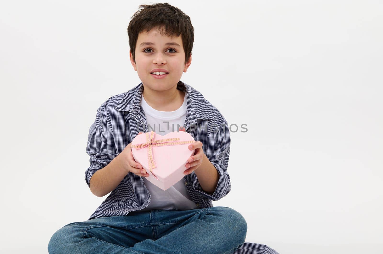 Handsome Hispanic boy, teenager, smiling with a toothy smile, showing a heart-shaped pink gift box with a happy surprise inside to the camera. Present for Valentine's Day, Mother's Day and Women's Day