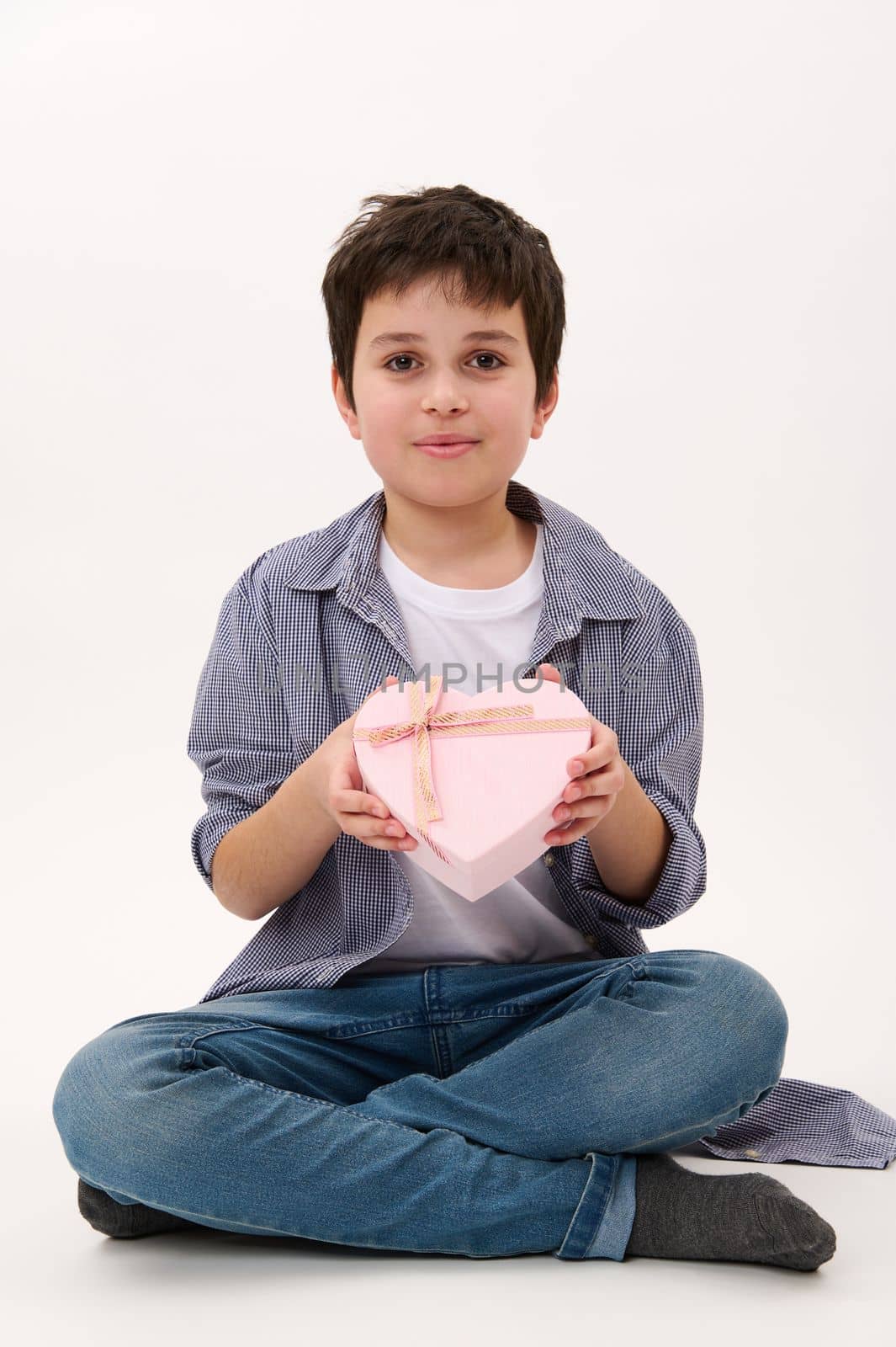 Vertical studio shot on white background, a ten year old handsome smiling Caucasian boy with a cute present for Valentine's Day, Mother's Day or Women's Day, in heart-shaped pink gift box. Copy space