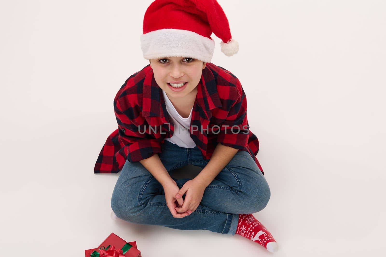 View from above of adorable multi-ethnic preteen boy in Santa hat and red plaid shirt, sitting on white background with christmas presents. Copy advertising space. Isolated portrait. New Year's event