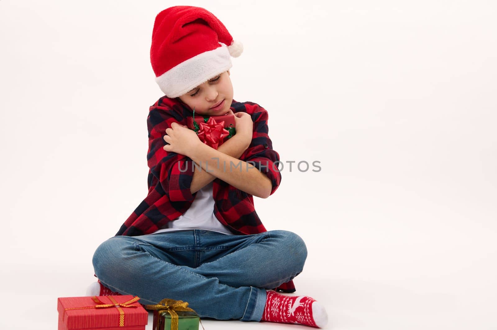 Adorable child boy in Santa hat, gently hugging a Christmas gift, isolated over white background, copy advertising space. Boxing Day. Happy festive atmosphere of winter holidays. New Year celebration