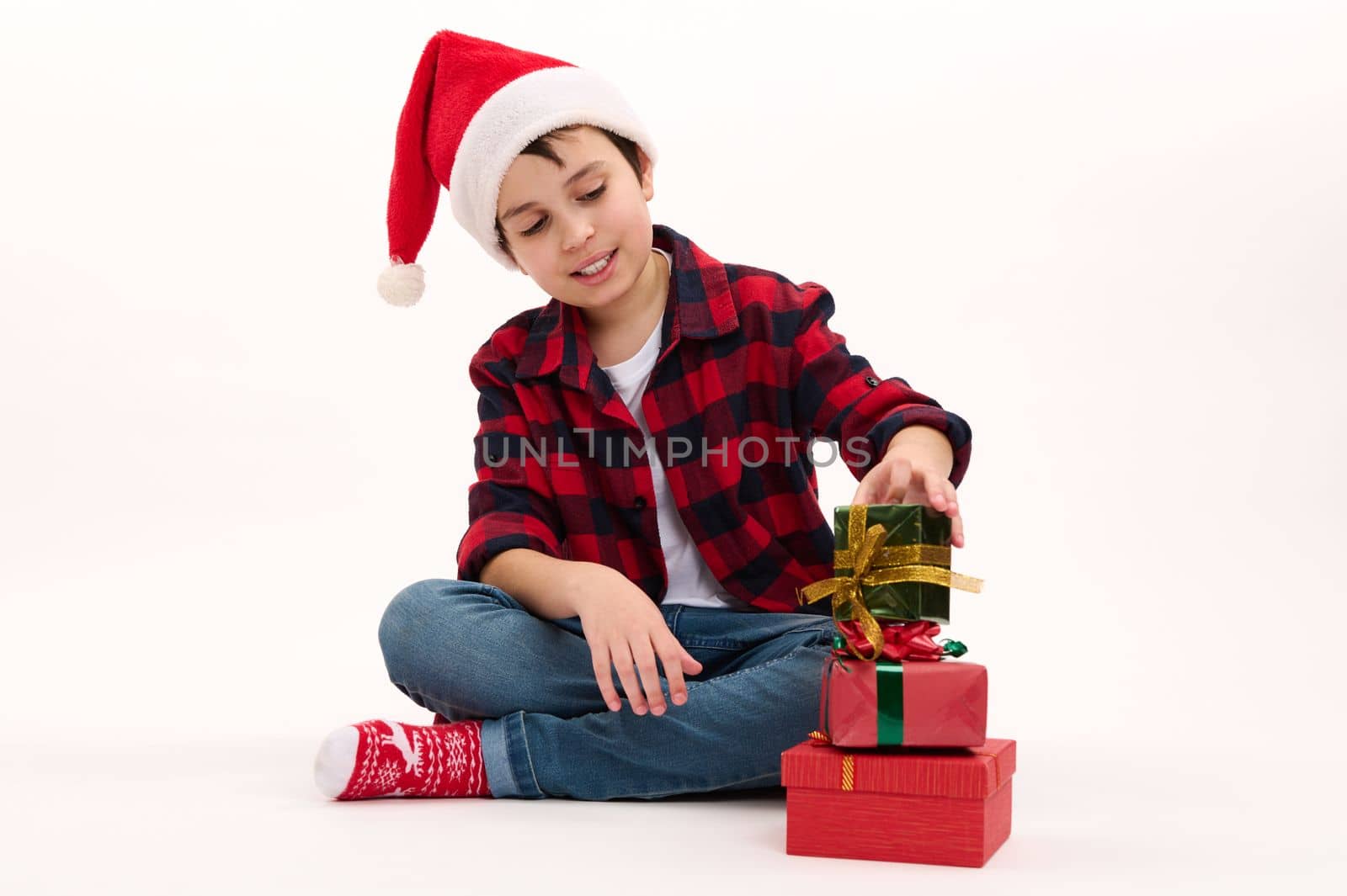 Portrait of a Caucasian lovely preteen boy wearing Santa hat, red plaid shirt, blue denim jeans and socks with Xmas pattern, stacking Christmas gifts, sitting on white background. New Year celebration